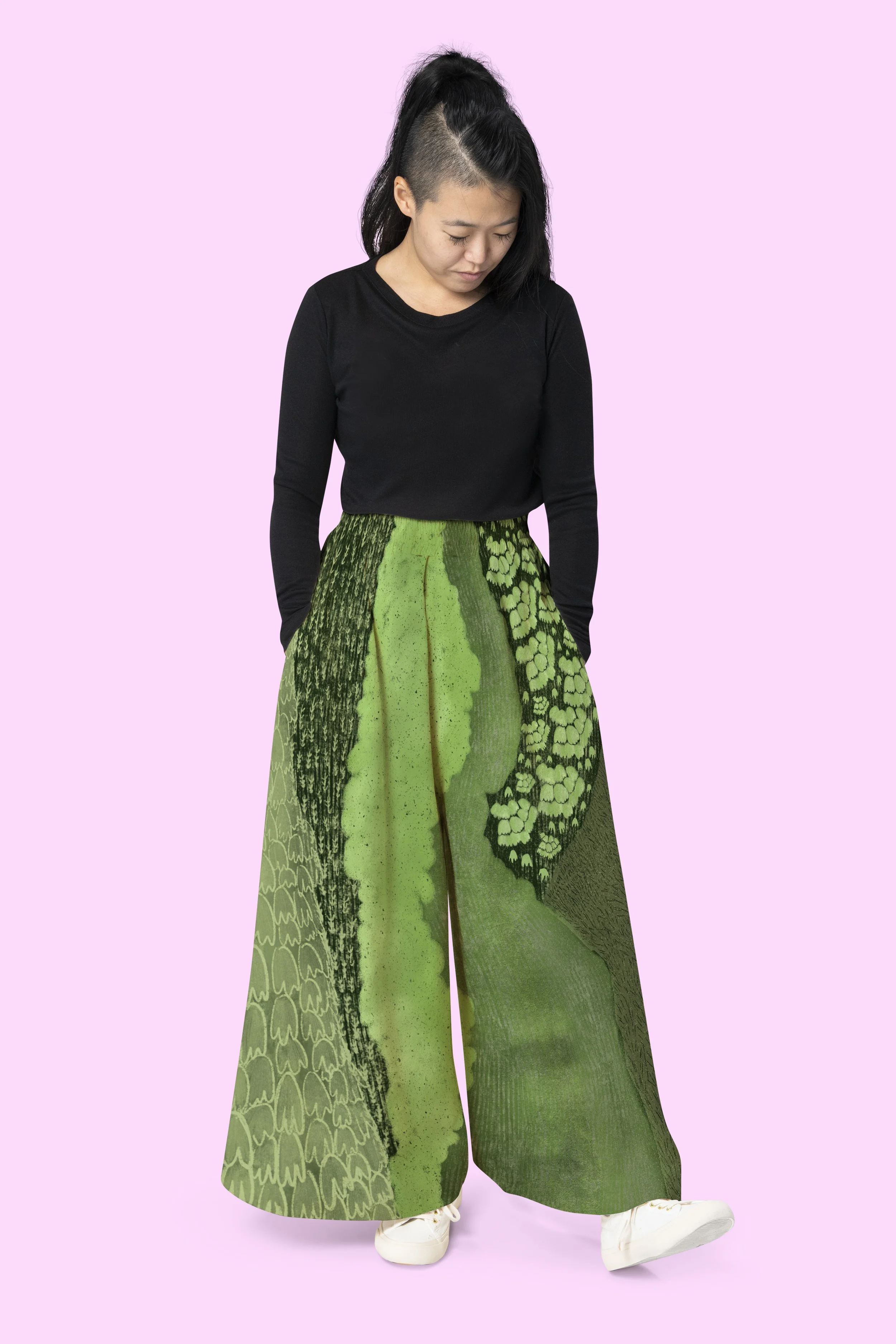 A person wearing a long green patterned skirt, a black long-sleeve shirt, and white sneakers, standing against a pink background.