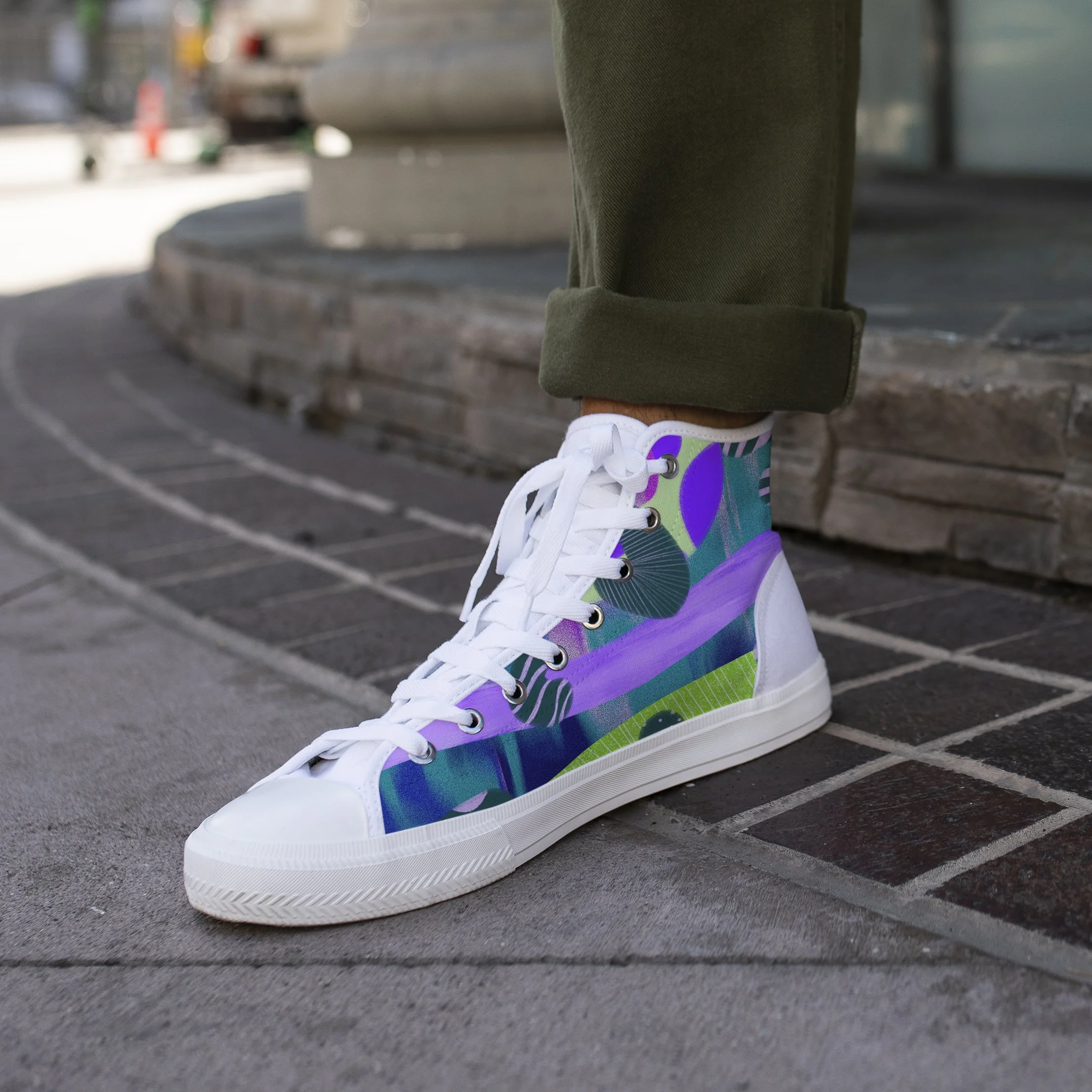 Close-up of a white sneaker with a colorful and patterned design, worn by a person standing on a stone-tiled sidewalk, with part of a green pant cuff visible.