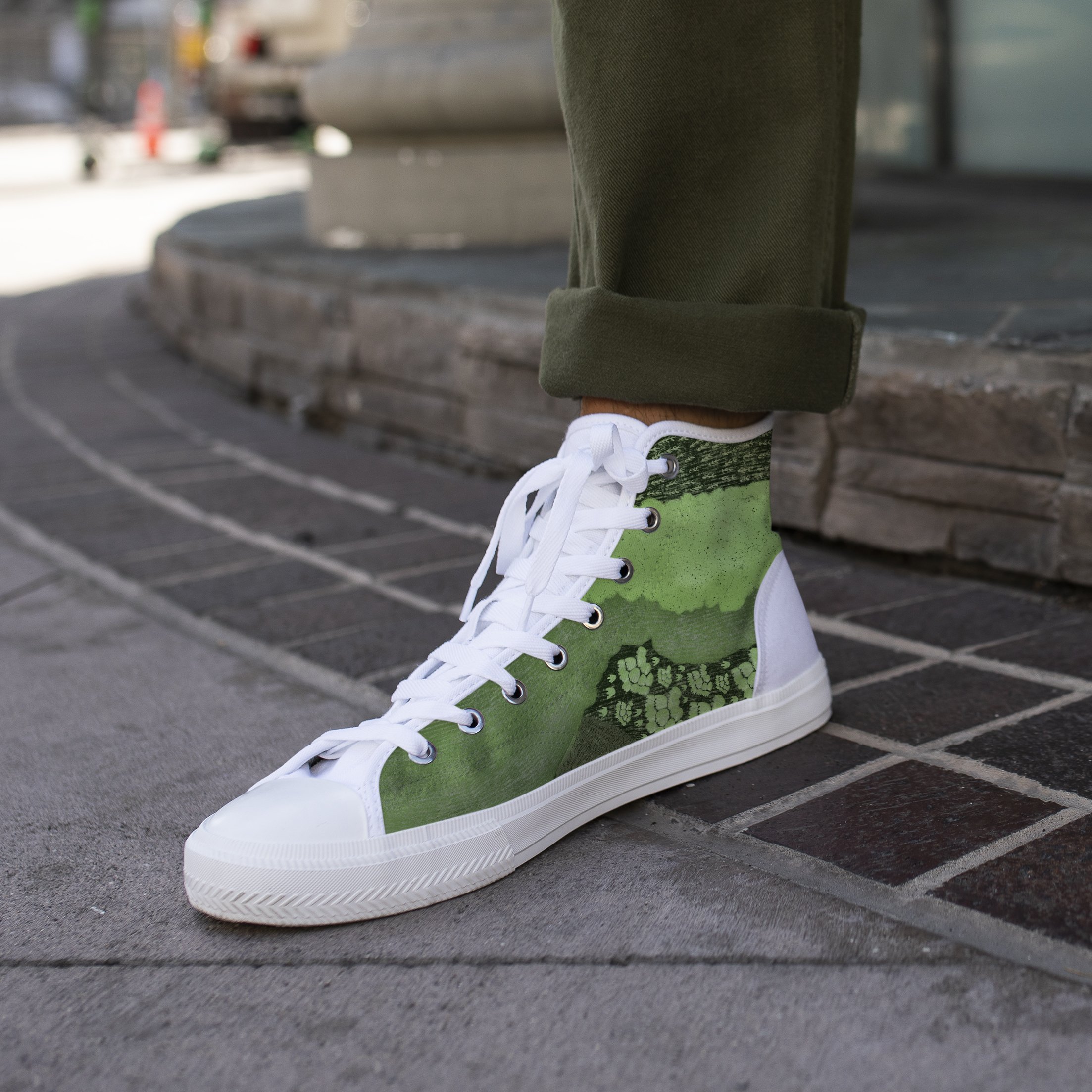 Close-up of a person's leg wearing green sneakers with white laces and white soles, rolled-up olive-green pants, standing on a paved area.