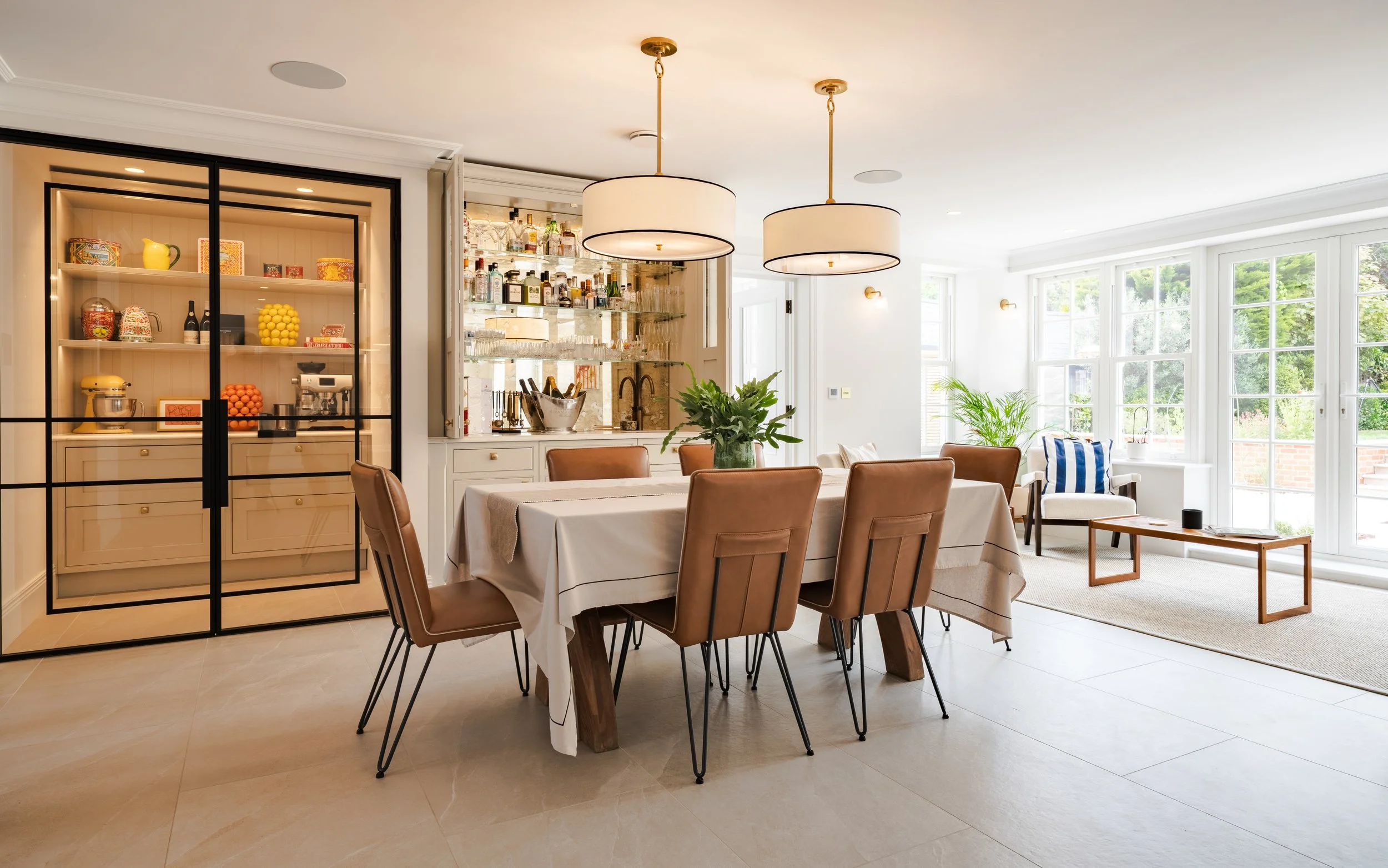 Bright dining room with a rectangular table, brown chairs, a vase of greenery, and a bar area with glass shelves filled with liquor bottles. Large windows with a view of trees and a small sitting area with a striped pillow and a small table.