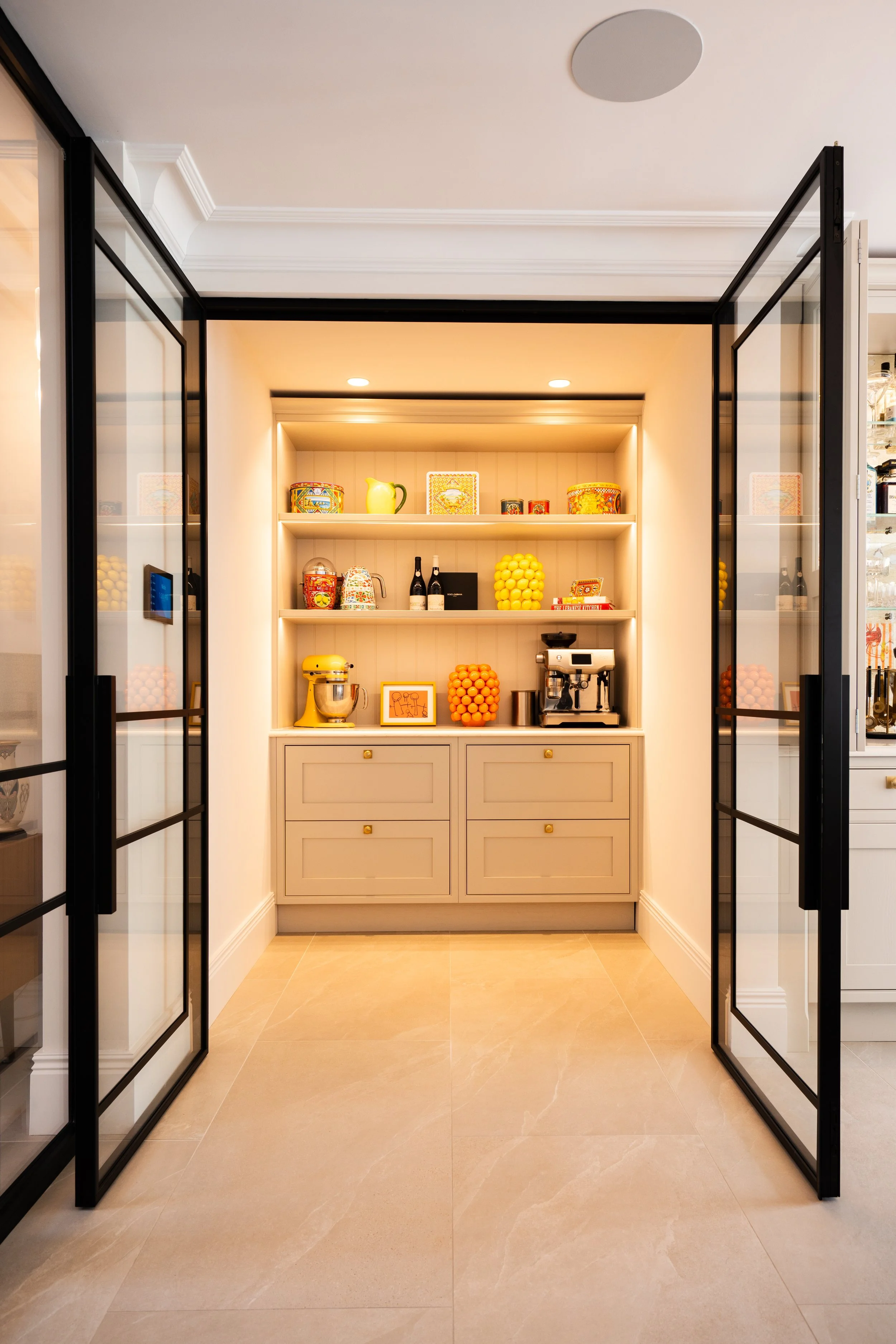 A built-in shelving unit with colorful decorative items, including ceramics, bottles, and artwork, situated between two black-framed glass doors in a modern interior space with beige tiled flooring.