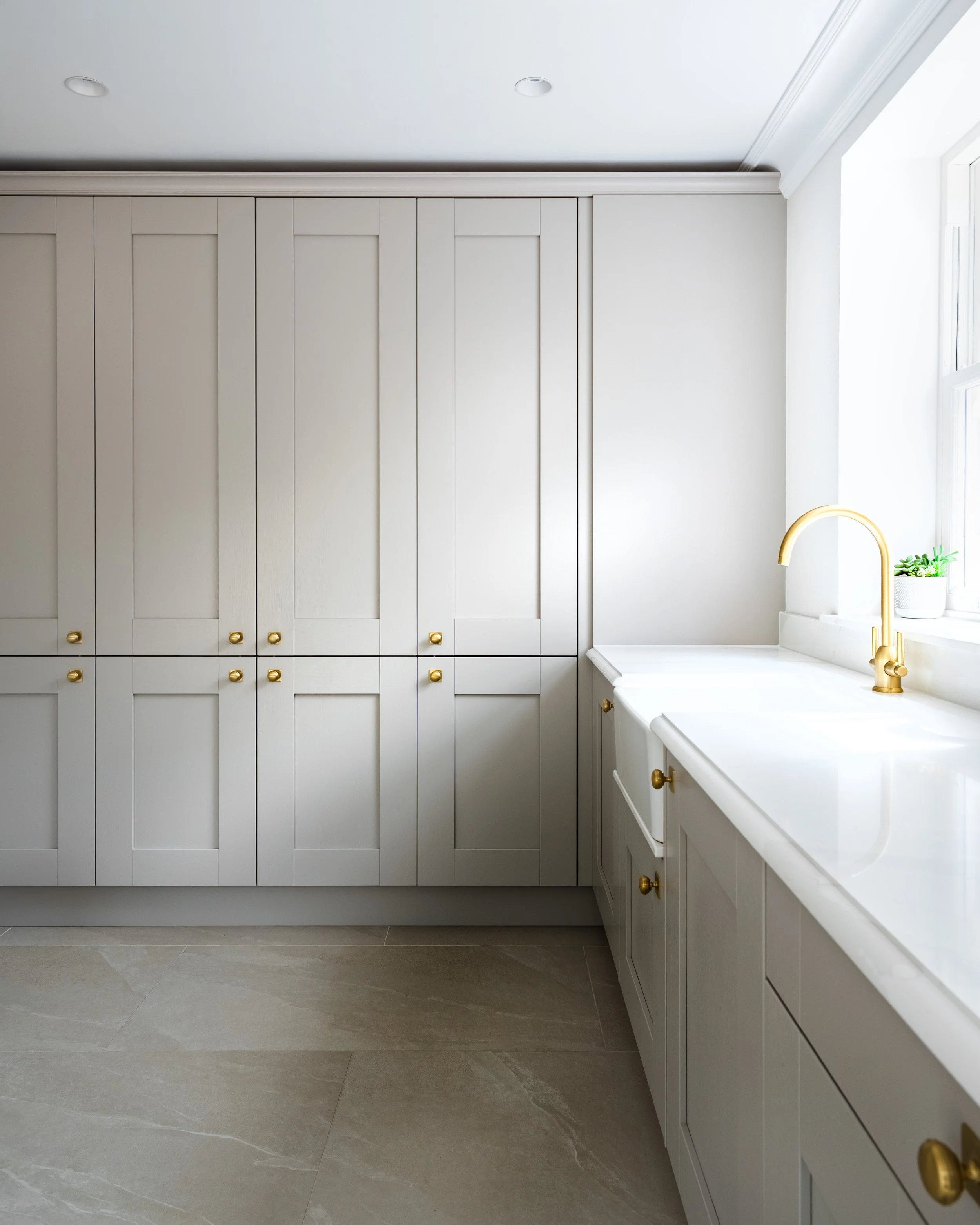 Modern kitchen with white cabinets with gold knobs, a white countertop, a gold faucet, and a small potted plant near the window.