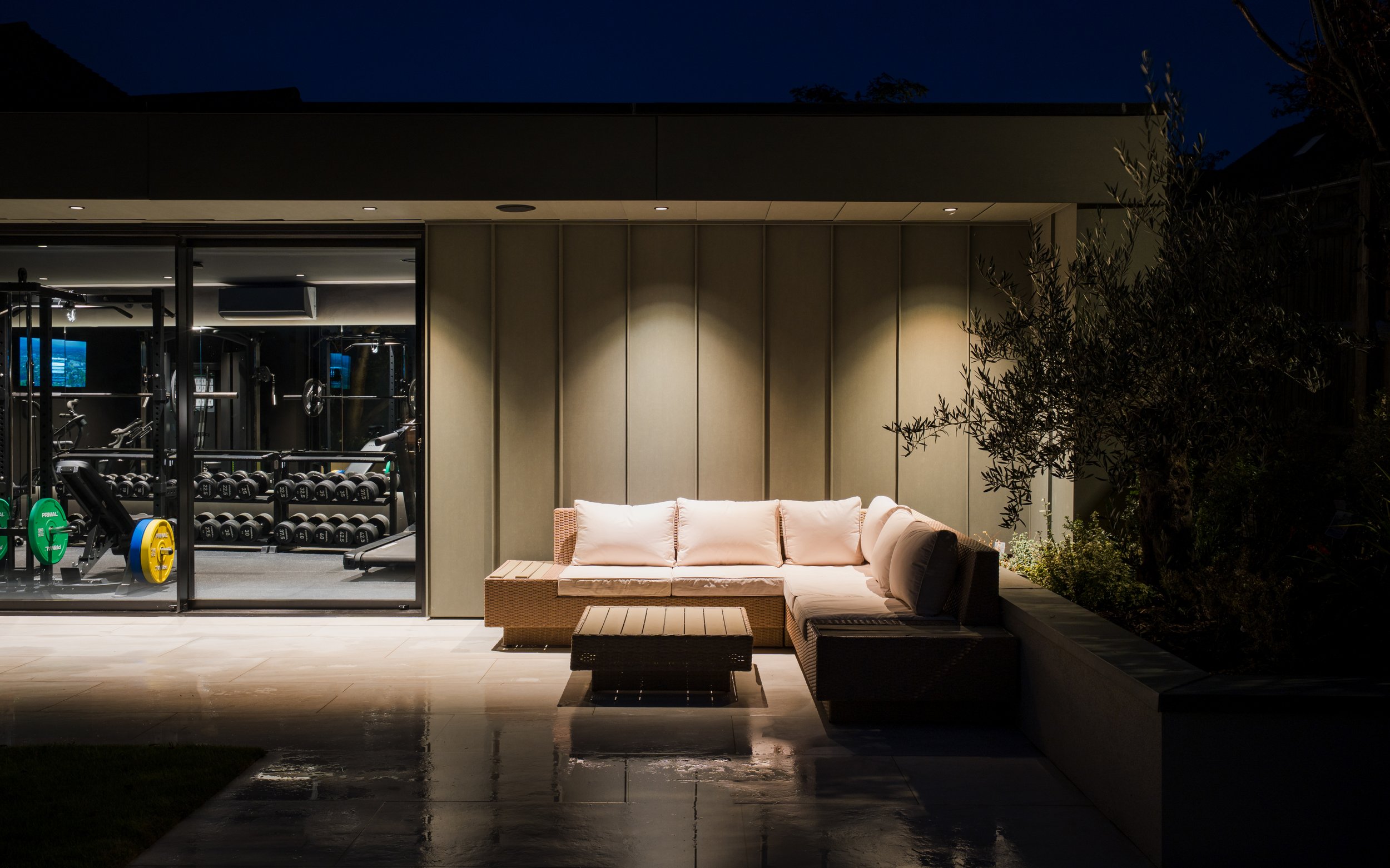 Night view of modern outdoor patio with a pink sectional sofa, small tables, garden plants, and a fitness gym visible through glass doors.