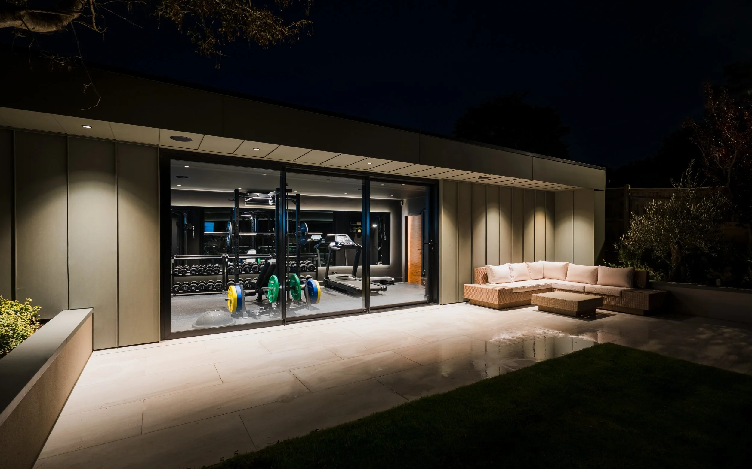 Nighttime view of a modern house with a glass wall showing a home gym with weights and exercise equipment, and an outdoor seating area with a beige sectional sofa and ottomans.