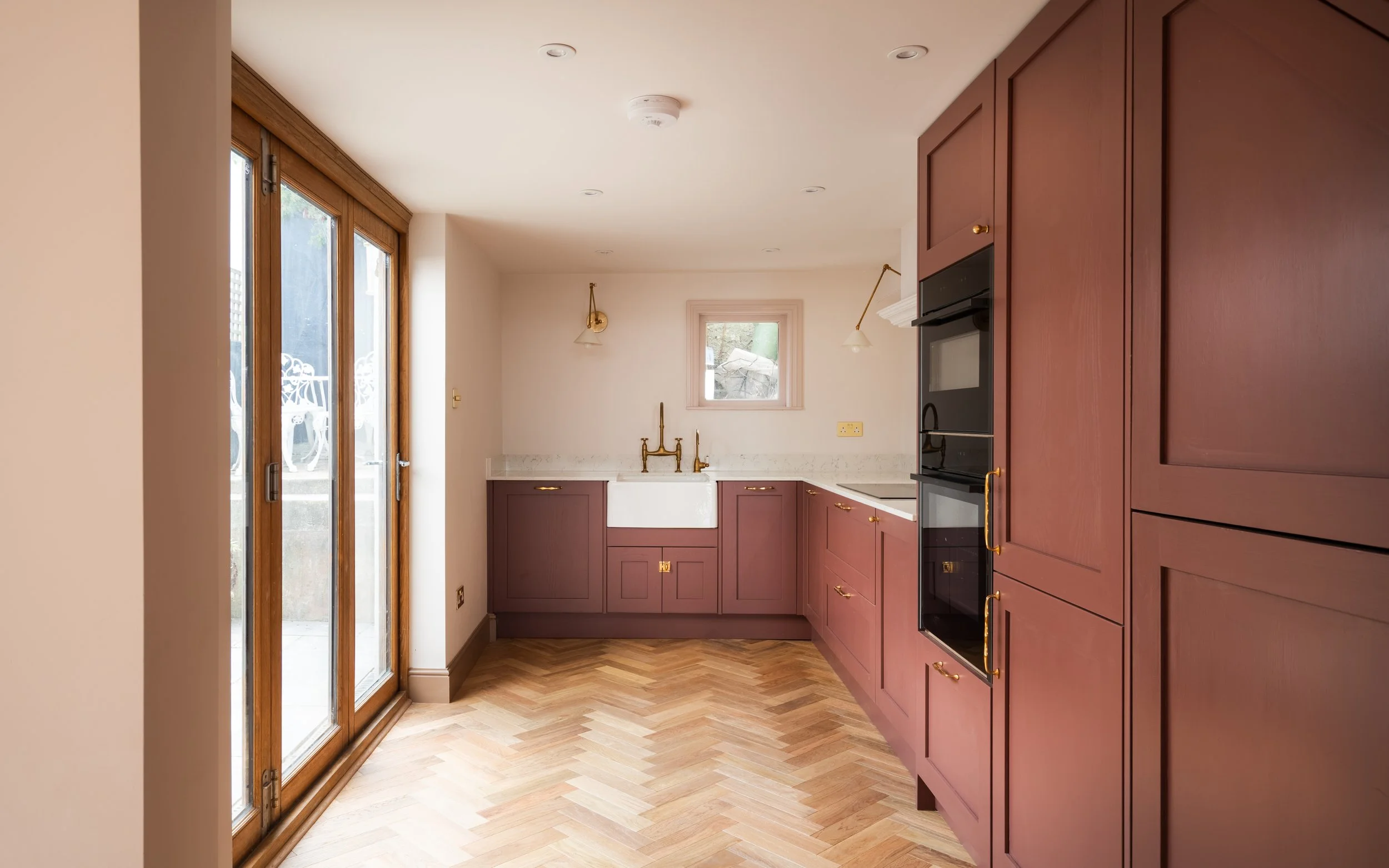 A kitchen with purple cabinets, a white farmhouse sink, gold fixtures, and wooden framed glass doors leading outside.