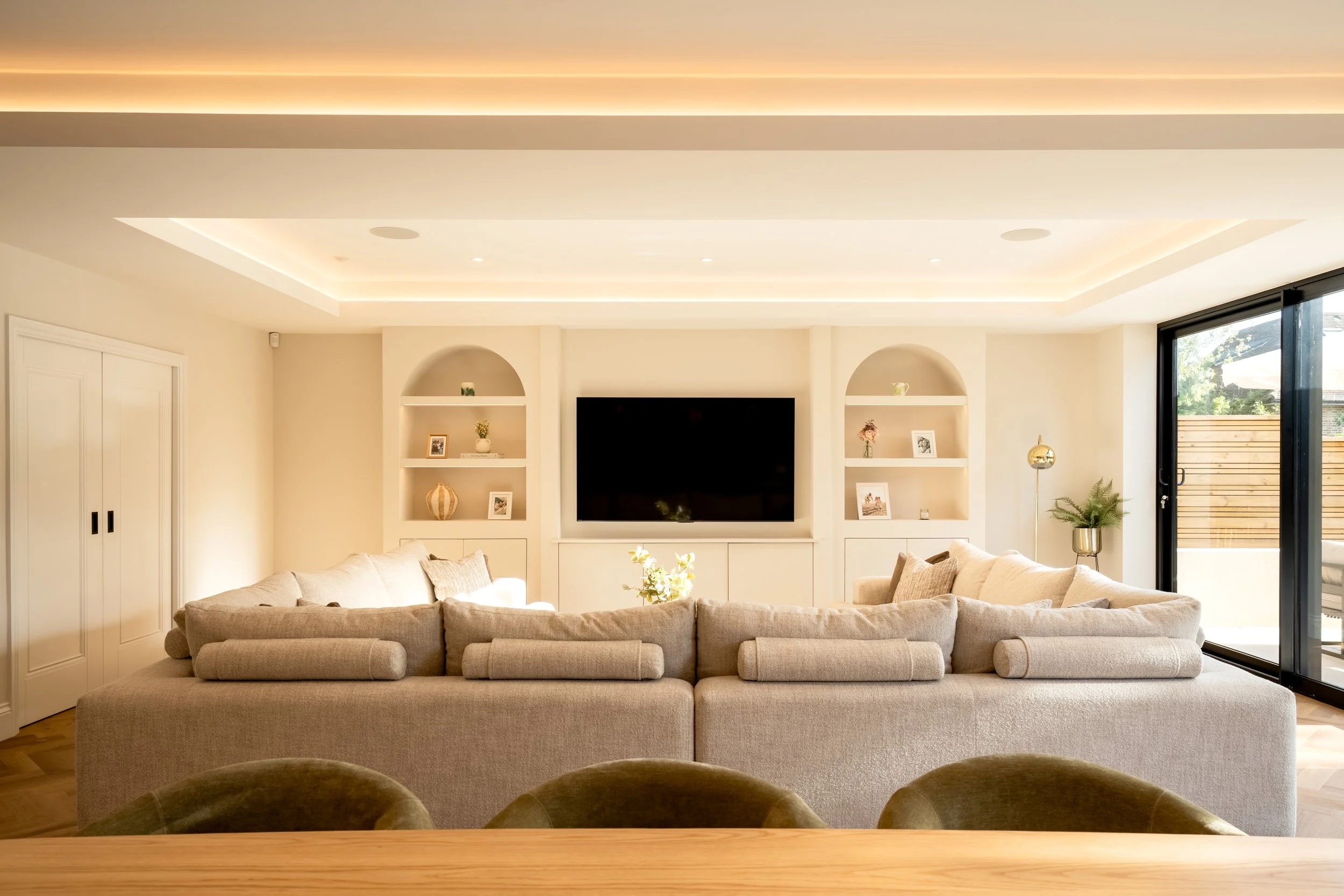 Modern living room with beige sectional sofa, wall-mounted flat-screen TV, built-in white shelves with decorative items, large windows with black sliding doors, and recessed ceiling lighting.
