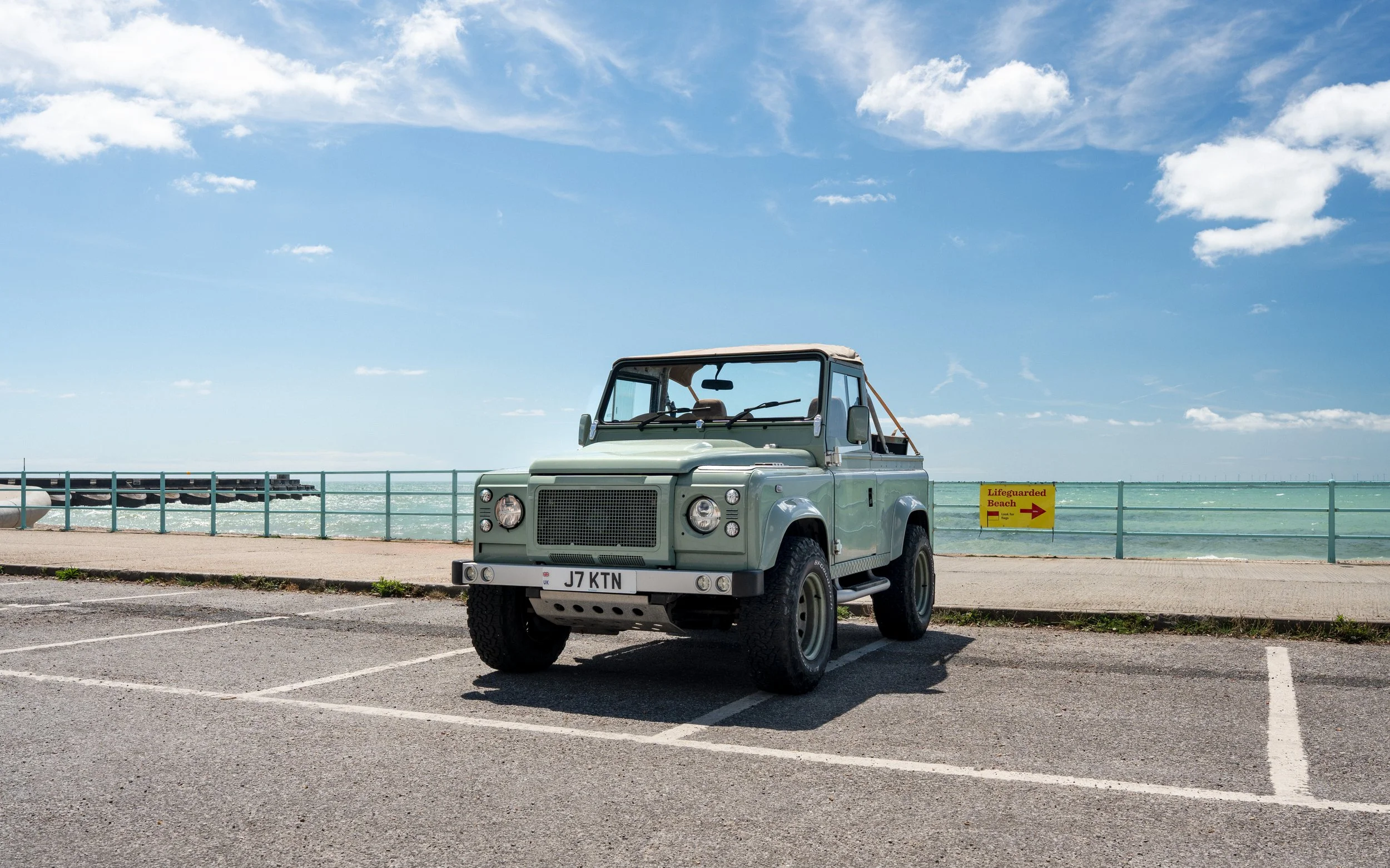 A light green Land Rover Defender parked near a beach with a sign indicating lifeguarded beach, blue sky with some clouds, and ocean in the background.