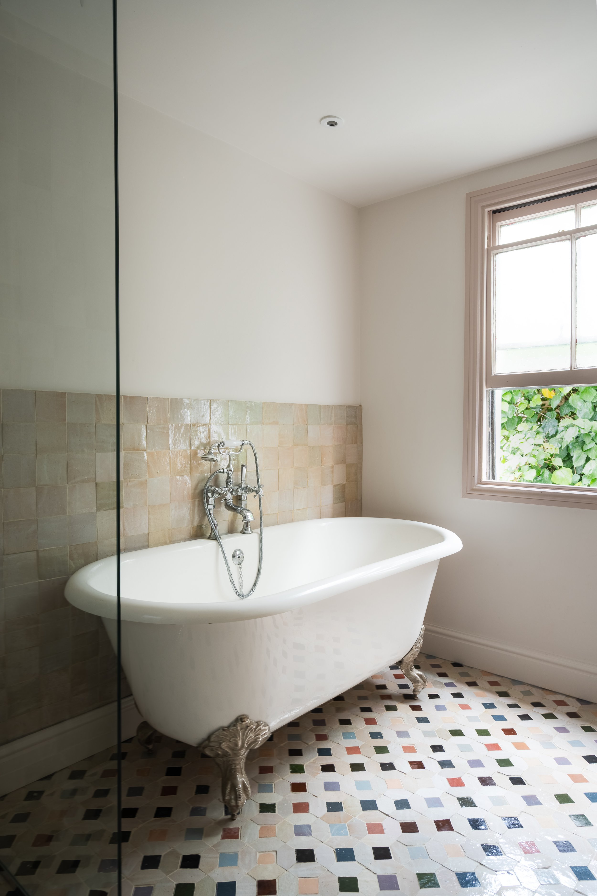 A vintage white clawfoot bathtub with ornate silver feet is in a bathroom next to a window with white trim. The wall behind the tub has beige tiles, and the floor has a mosaic pattern with small square tiles in various colors. The bathroom has white 