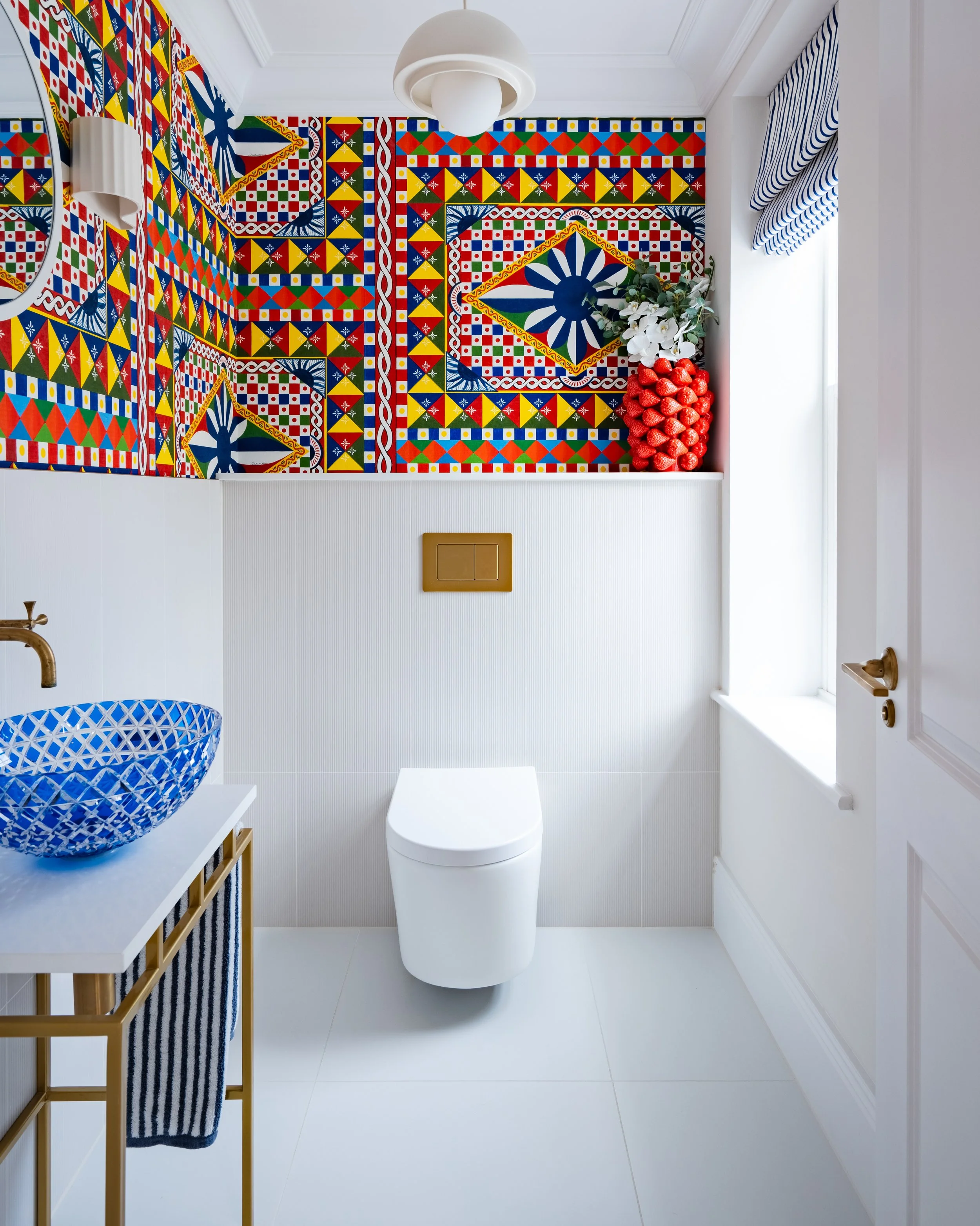 A modern, minimalist bathroom featuring a colorful and patterned tiled wall behind a white toilet. The bathroom has a white vanity with a blue patterned ceramic sink, a gold-framed mirror, a white window with striped blue and white curtains, a white 