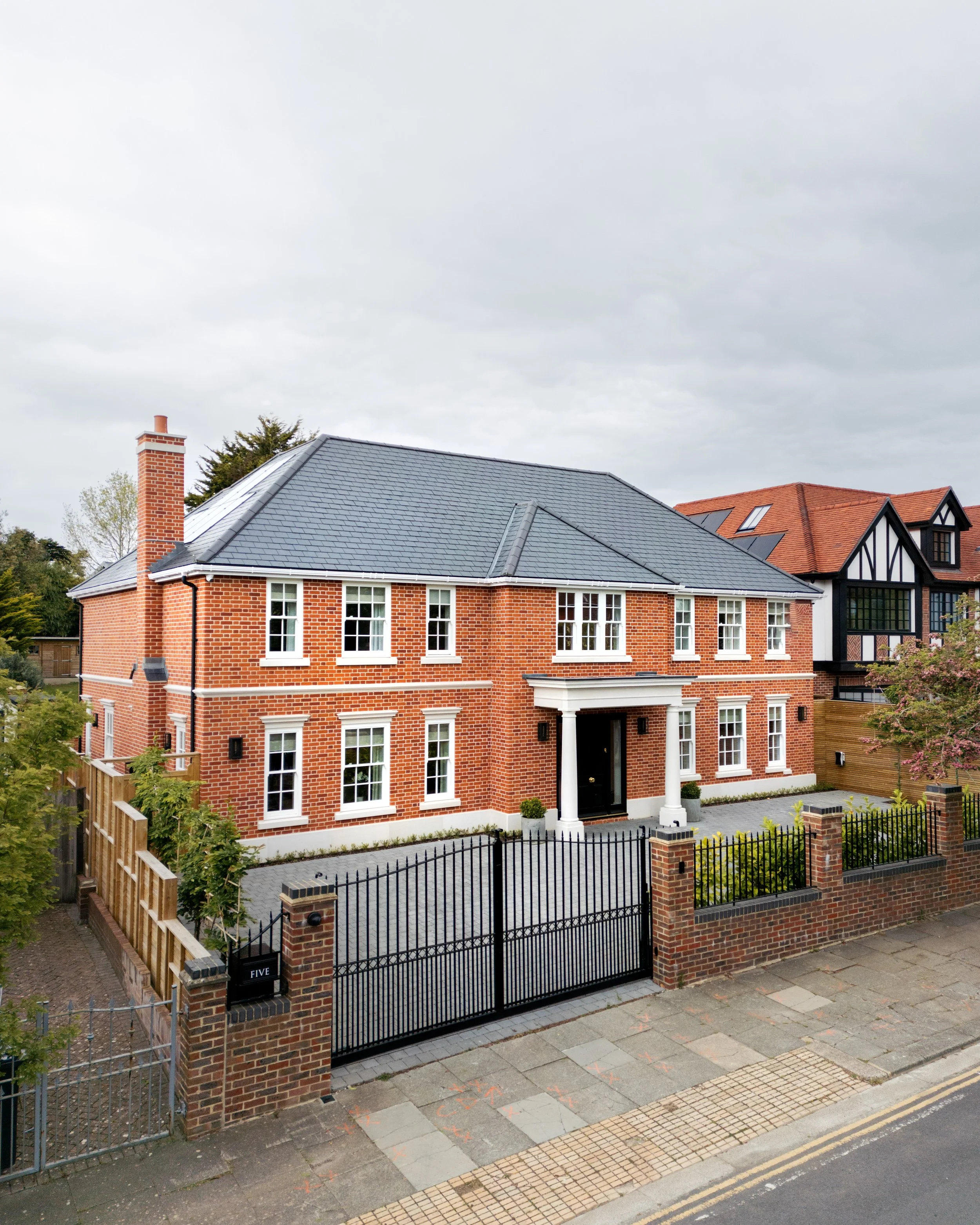 A large brick house with a dark roof, white-framed windows, and a black front door. The house is surrounded by a brick and iron gate and fence. There is a paved path leading to the entrance, and a sidewalk with street markings in front.