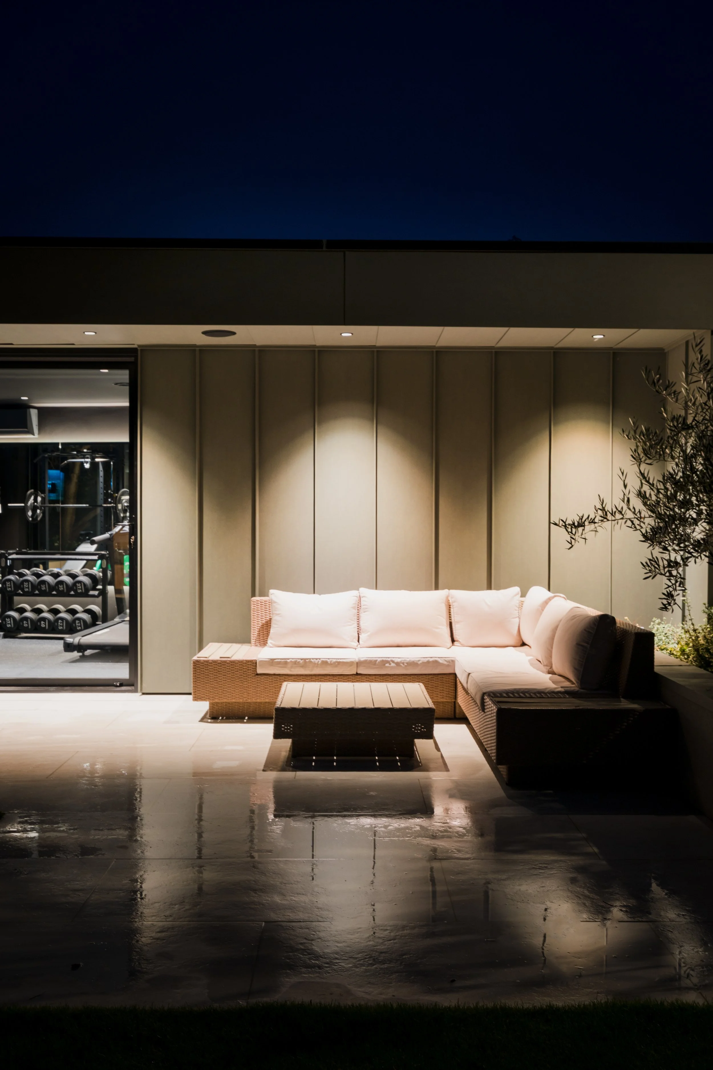 Nighttime outdoor patio with beige sectional sofa, coffee table, and a tree with small leaves.