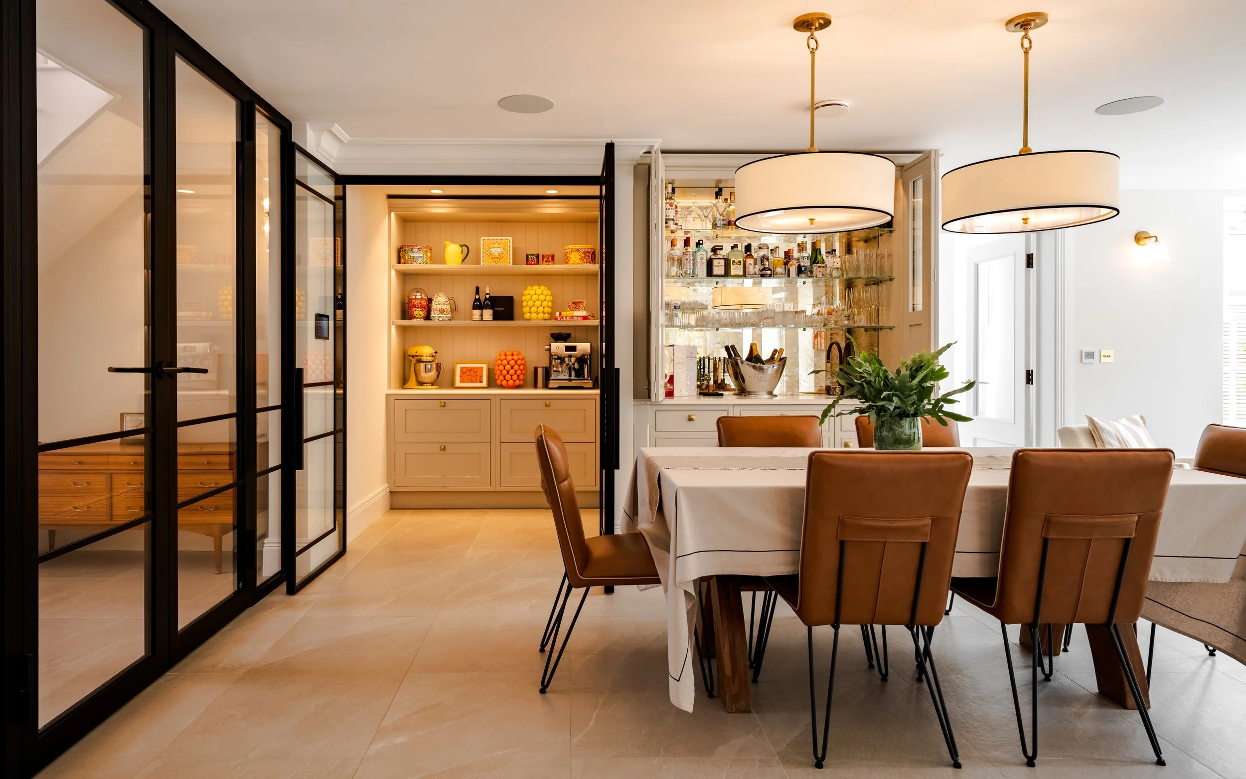 A dining area with a large table covered with a white tablecloth, surrounded by brown leather chairs, a potted plant in the center, and large circular pendant lights hanging from the ceiling. There is a built-in bar with bottles and glasses on shelve