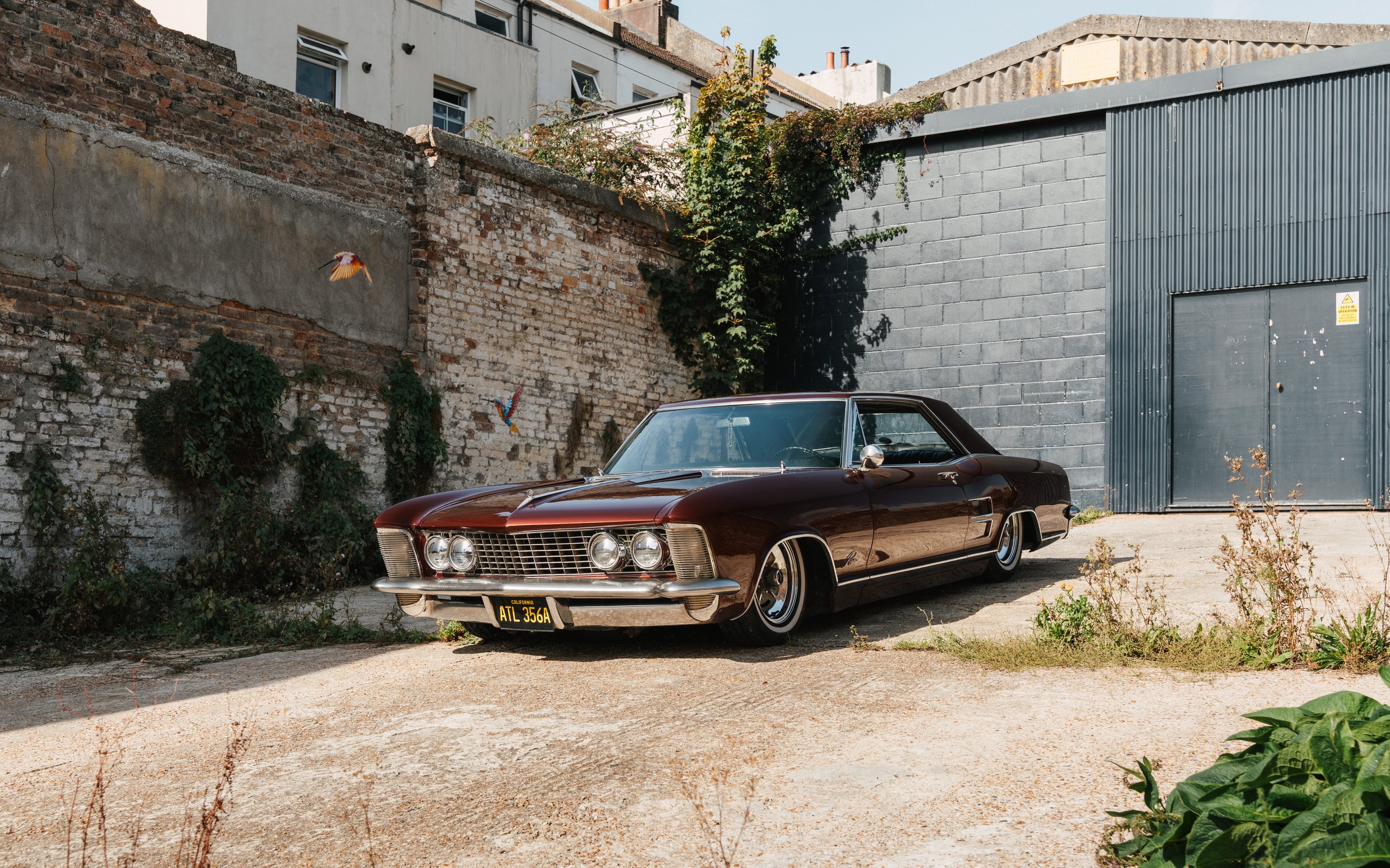 A vintage maroon car parked in a gritty urban lot with a brick wall and a gray industrial building in the background. The car has a yellow California license plate with the characters ATL 356A.