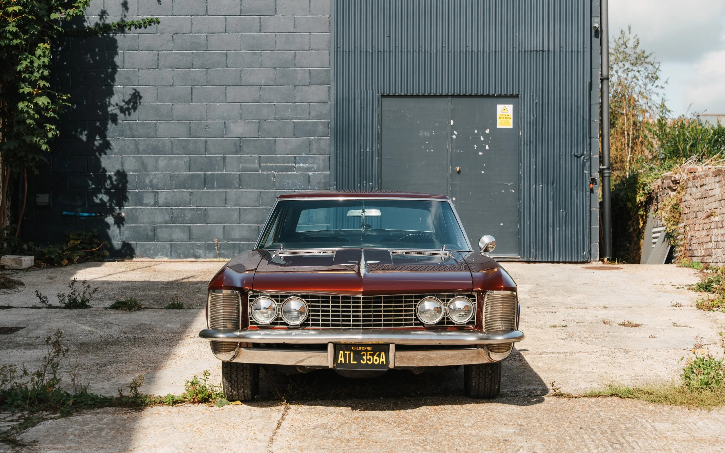 Vintage maroon car with California license plate ATL 356A parked in front of a gray cinder block wall and a metal door, with greenery on the sides.