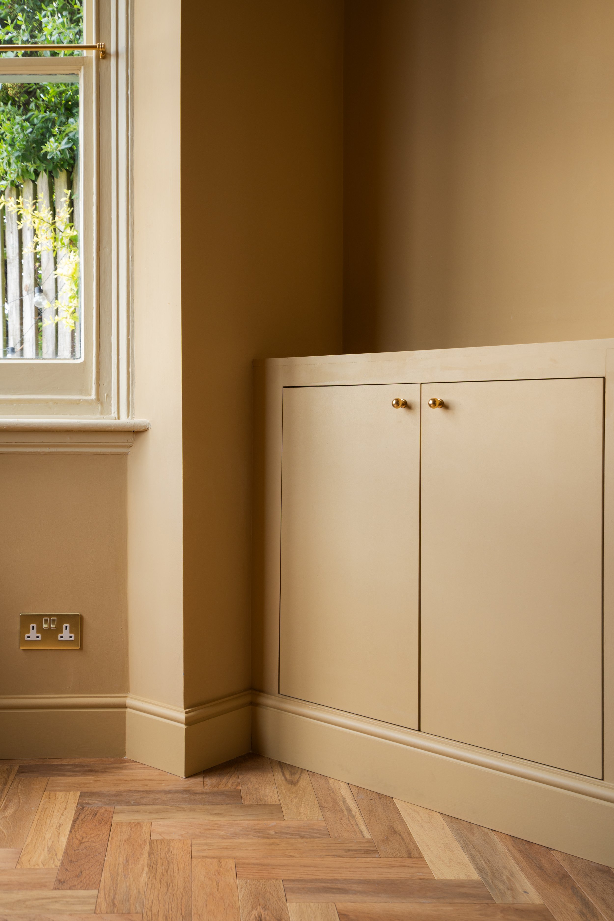 Corner of a room showing a window with a garden outside, a cream-colored cabinet with gold knobs, a beige wall, and a wooden parquet floor.