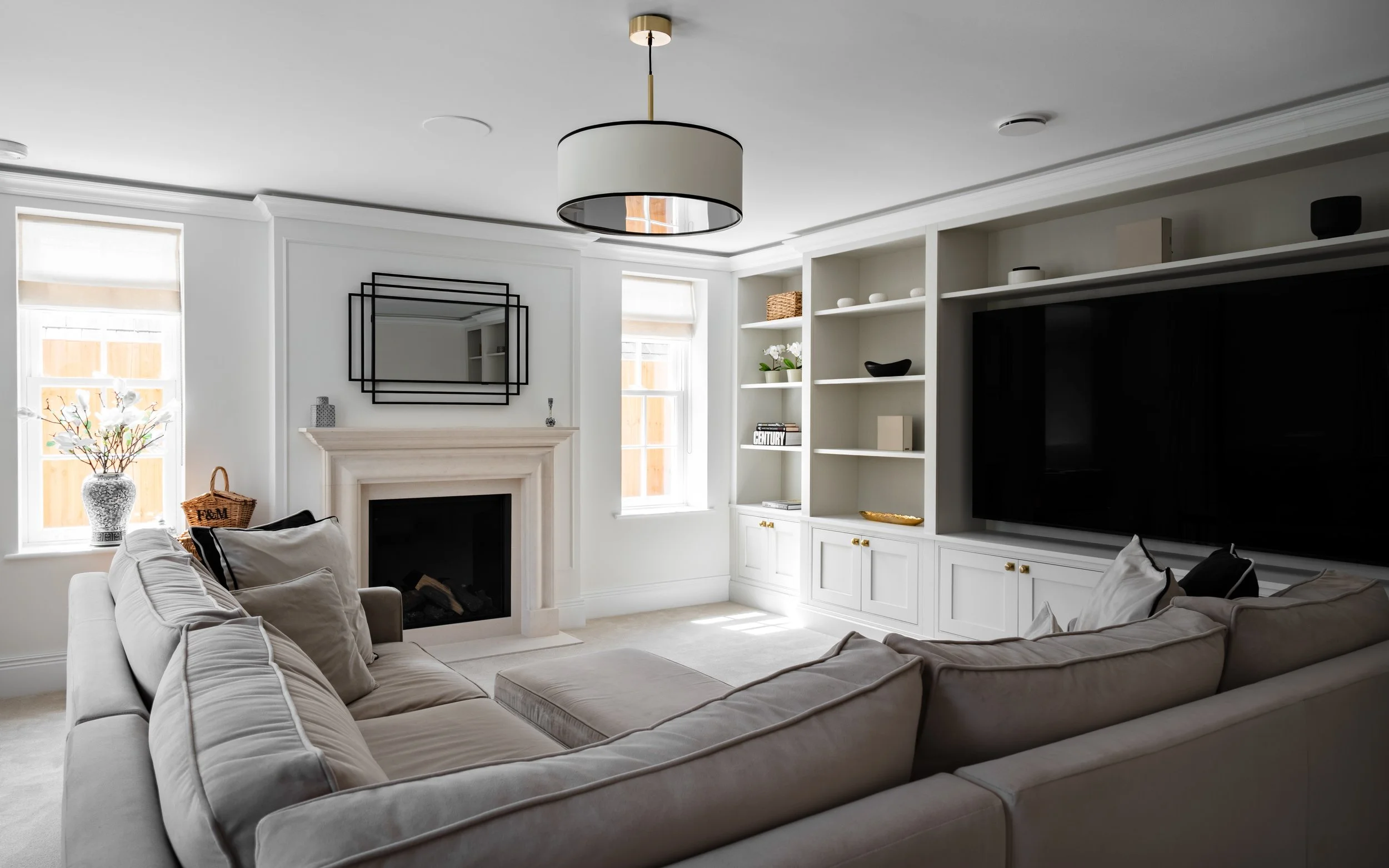 A modern living room with a circular ceiling light, white walls, and a beige sofa with pillows. There is a fireplace with a mantel, two windows with shades, and built-in white shelving with decorative items and books. A large, flat-screen TV is mount