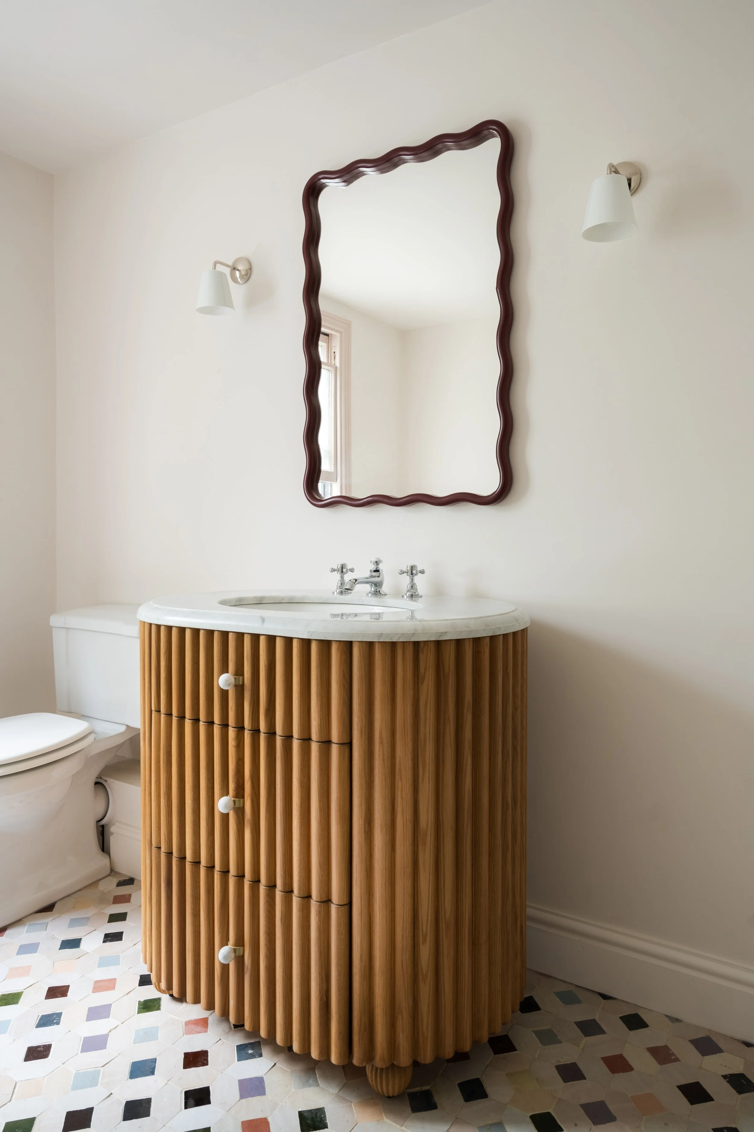 A bathroom vanity with a wooden cabinet featuring vertical slats, marble countertop with a built-in sink, and two cabinet knobs. Above the vanity is an irregularly shaped mirror with a wavy dark frame. Two white wall sconces are mounted on either sid