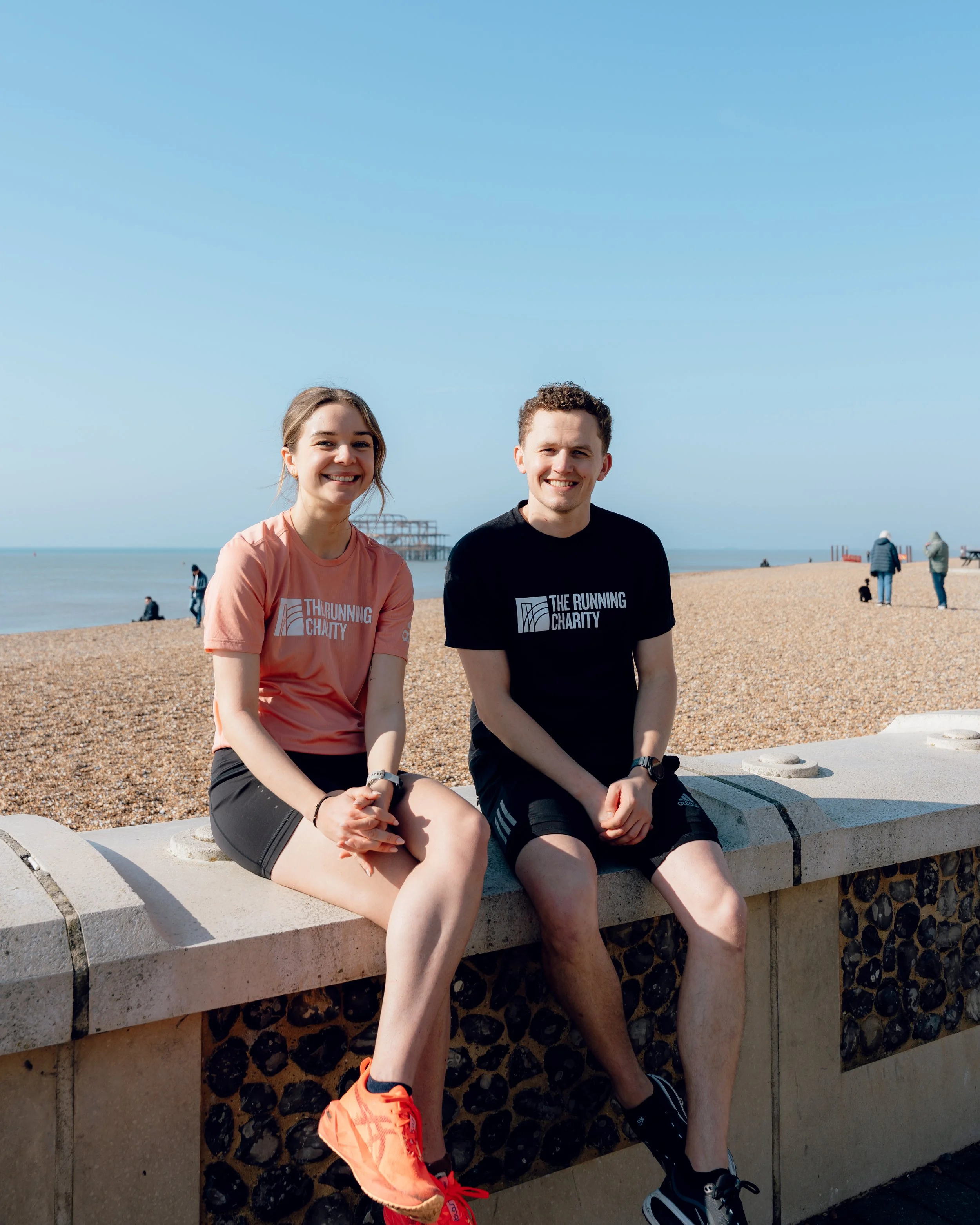 A man and a woman sitting on a concrete bench at the beach, smiling at the camera. The woman is wearing a pink T-shirt, black shorts, and orange running shoes, and the man is wearing a black T-shirt, black shorts, and black running shoes. In the back