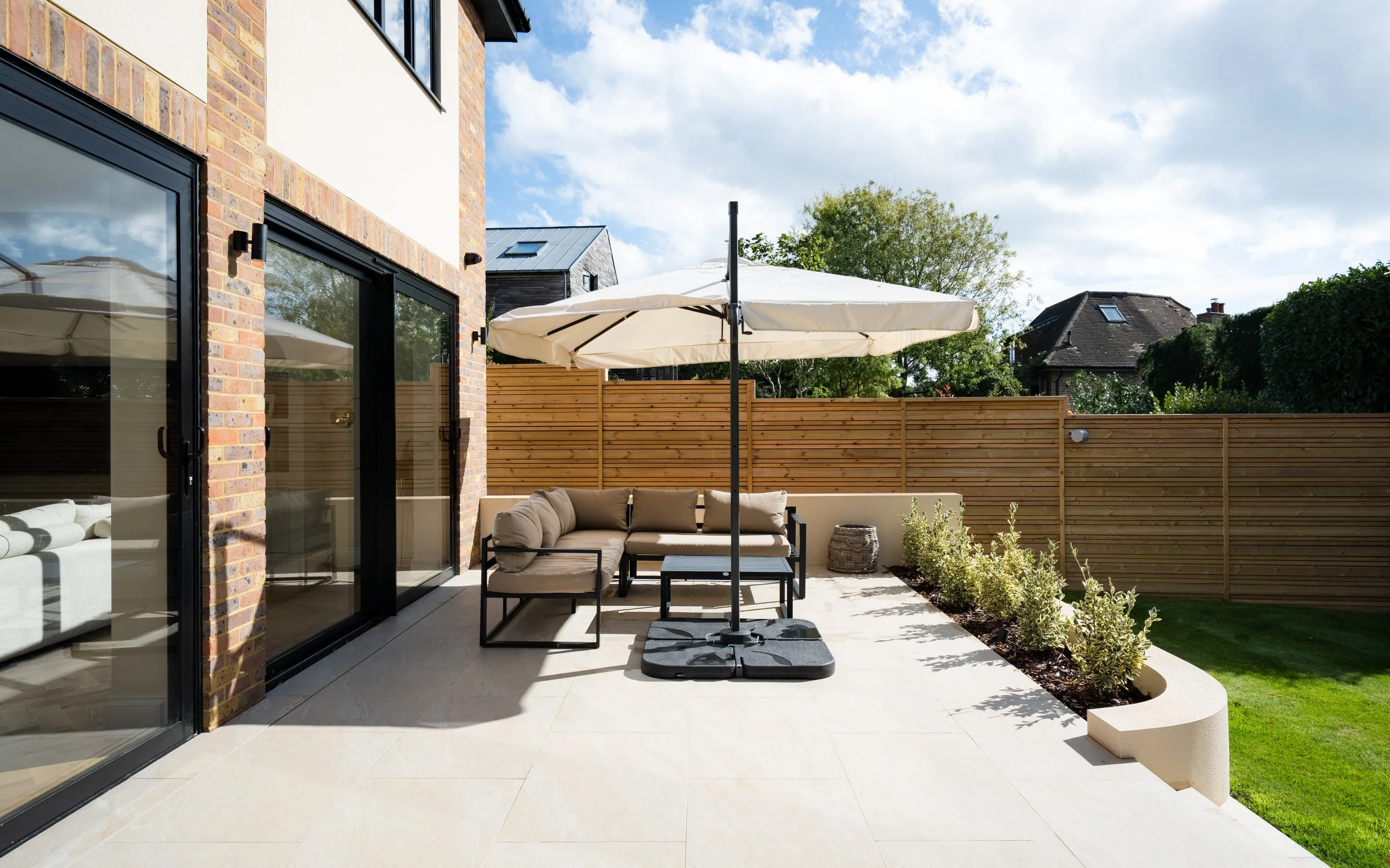 A modern backyard patio with outdoor furniture, a large white umbrella, a wooden privacy fence, and a small garden area with plants and grass. The house has large glass sliding doors and brick and white walls, under a partly cloudy sky.