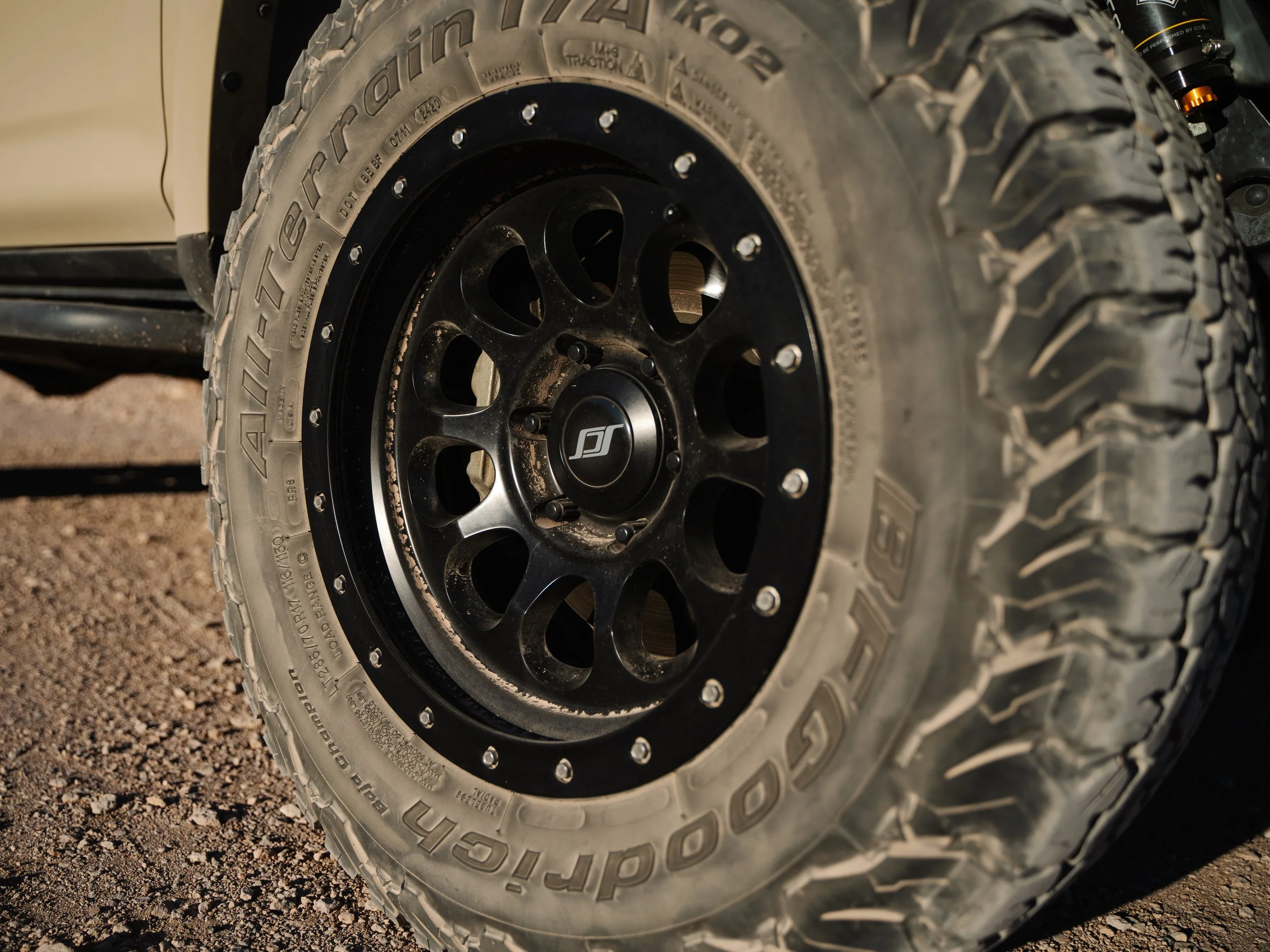 Close-up of an off-road tire mounted on a black rim on a vehicle, with visible dirt and gravel underneath.