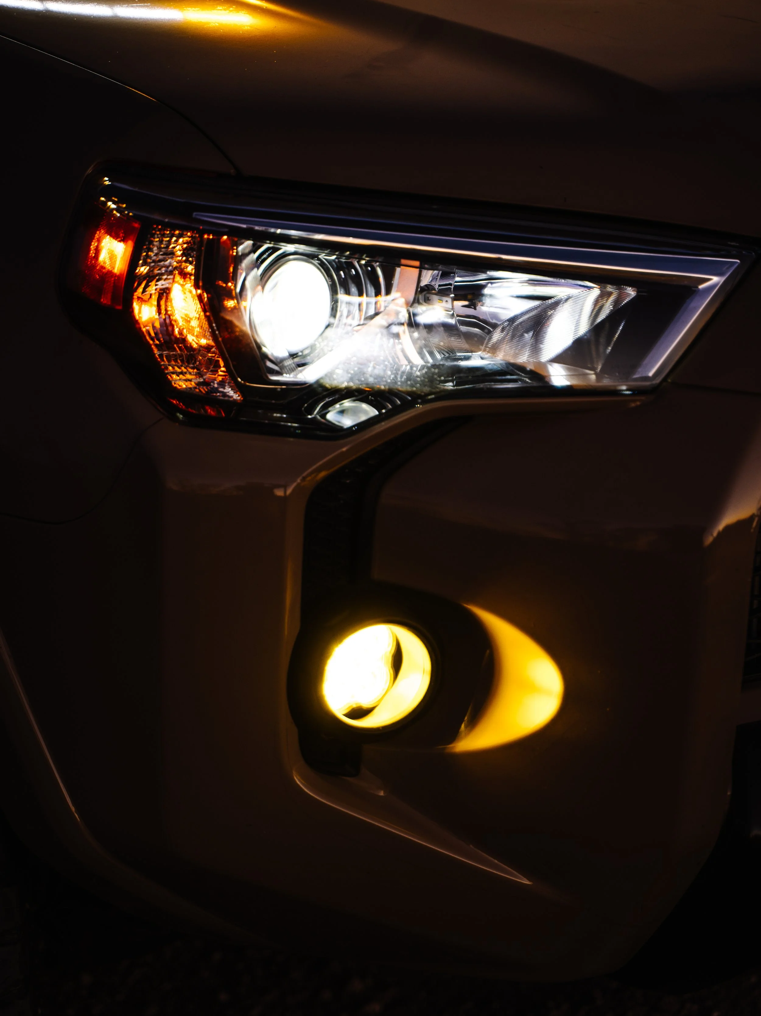 Close-up of a car's front headlight and fog light turned on at night.