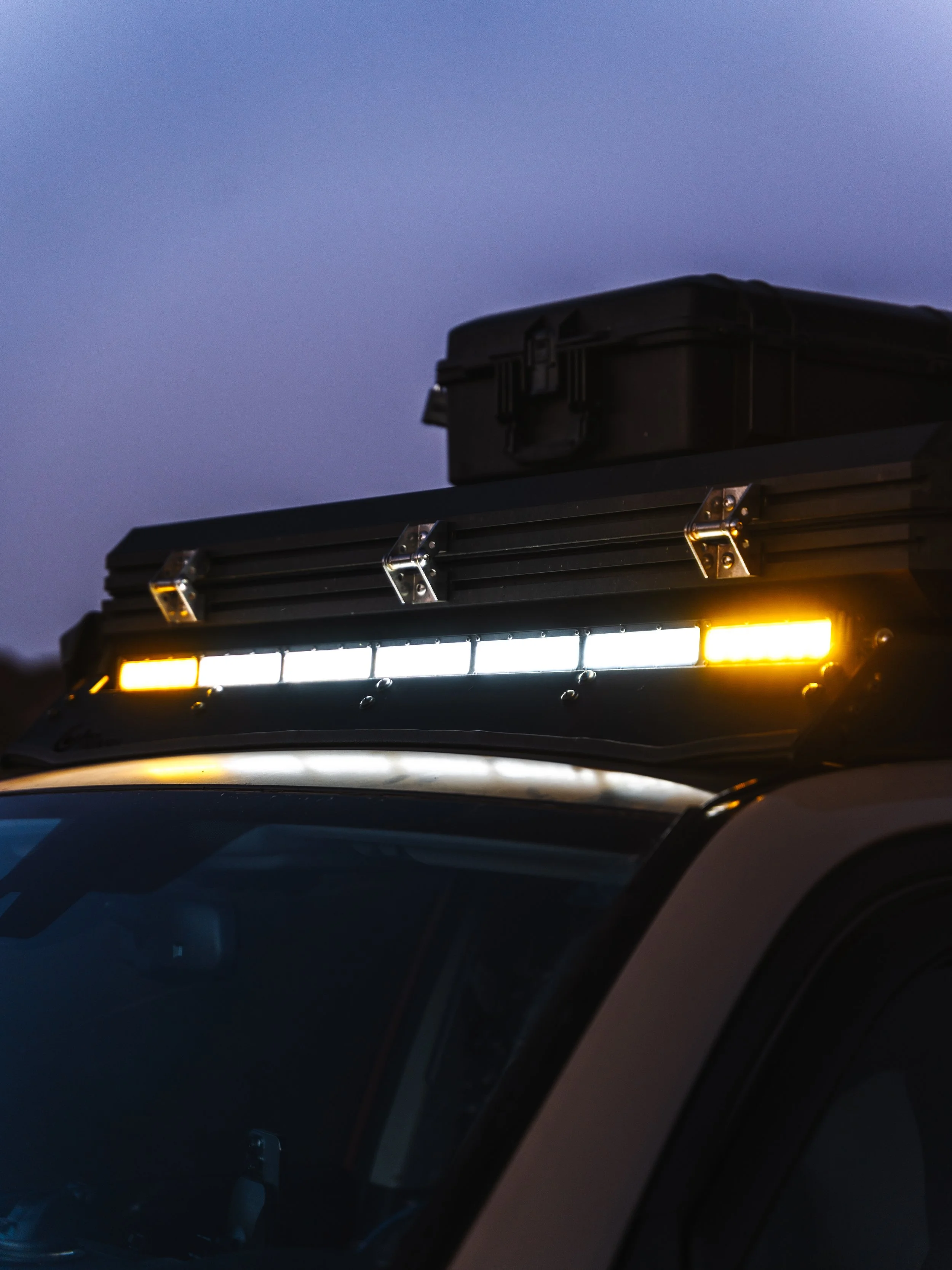 Close-up of the roof of a police vehicle during twilight, showing LED light bars in white and yellow, with a black storage box on top.