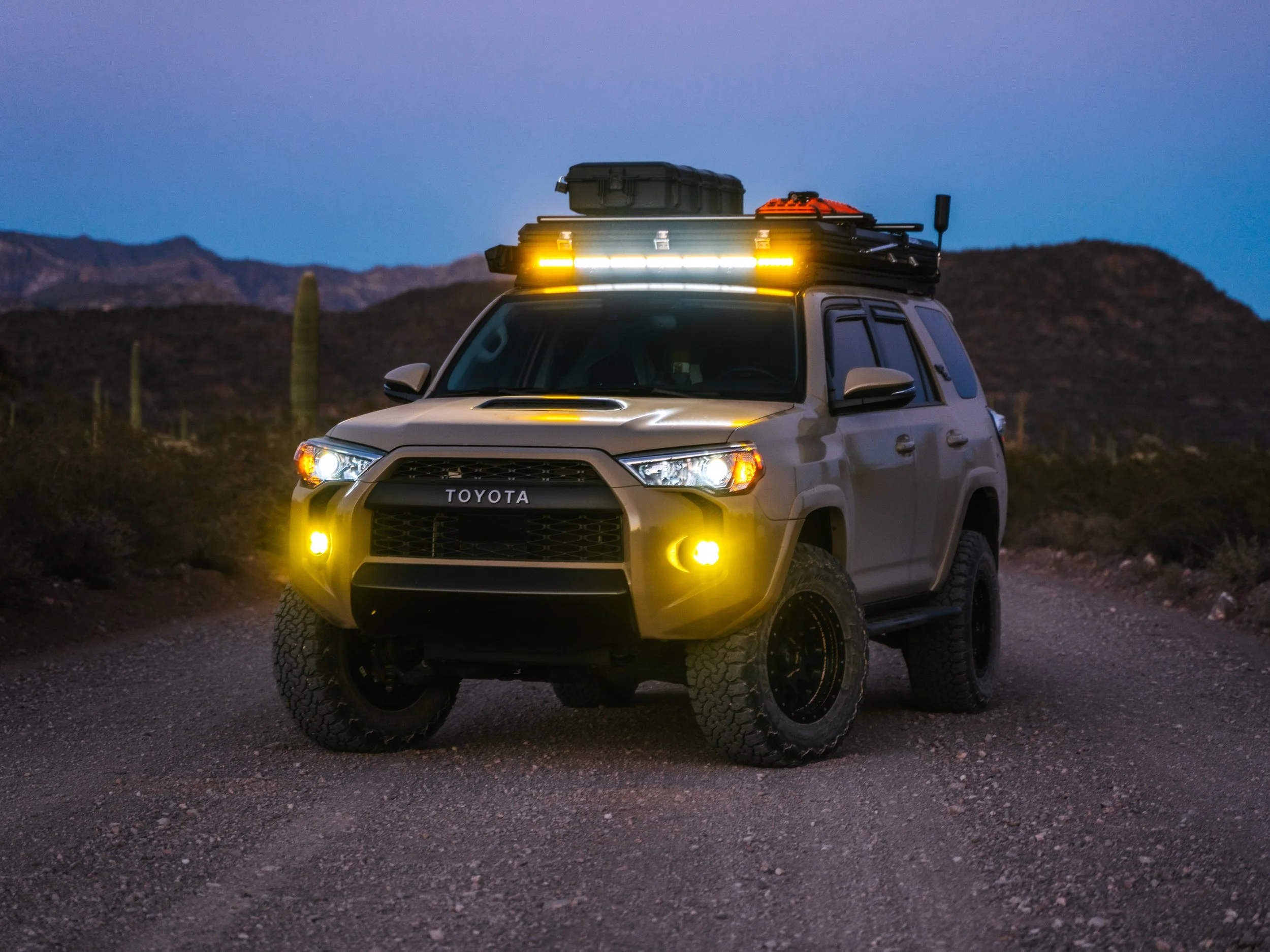 A beige Toyota off-road vehicle with yellow headlights and roof lights parked on a desert trail at dusk, with mountains and cacti in the background.