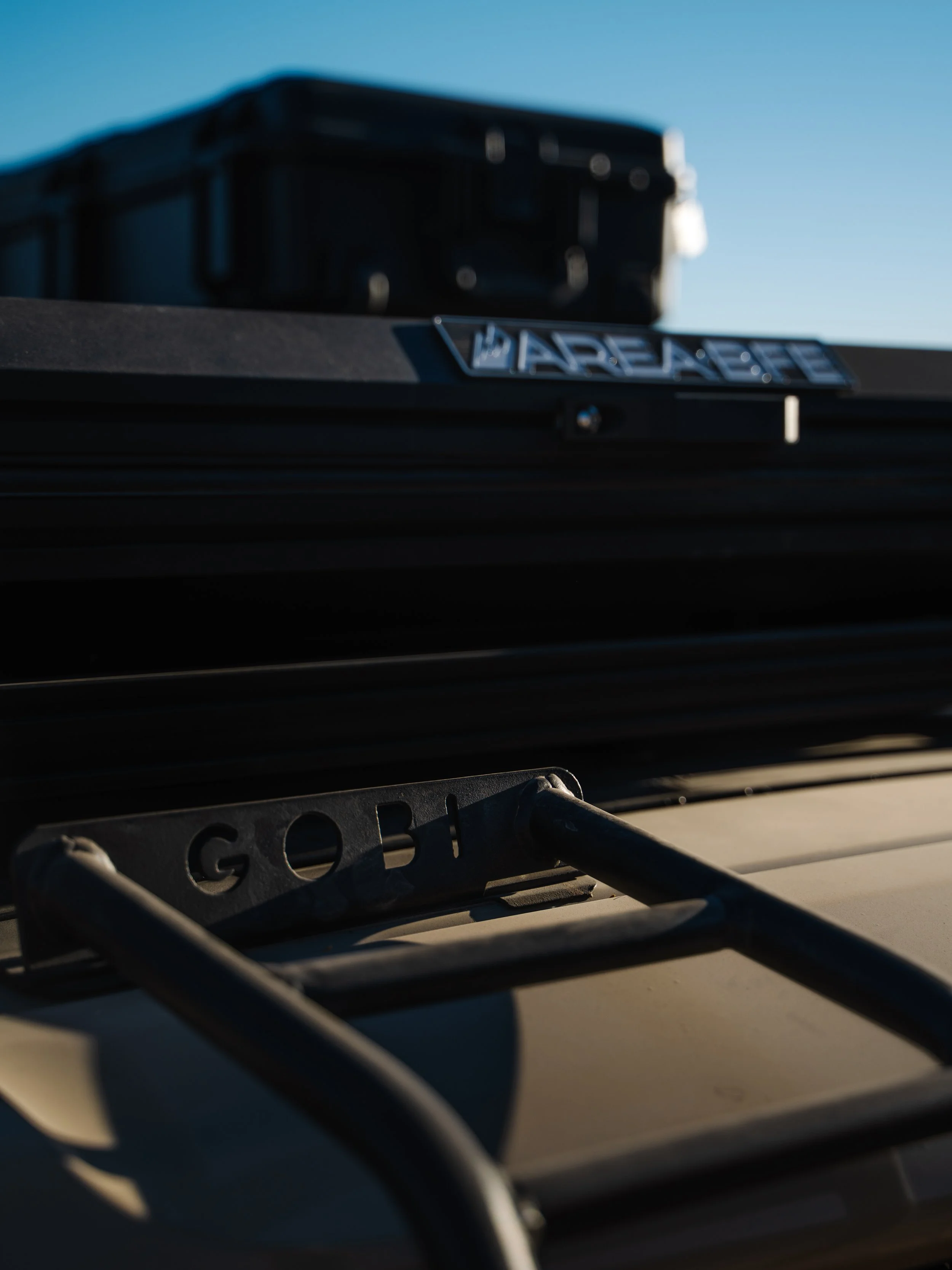 Close-up of a car roof with a roof rack labeled 'Gobi', and a black storage box labeled 'Arbortech' mounted on top, against a clear blue sky.