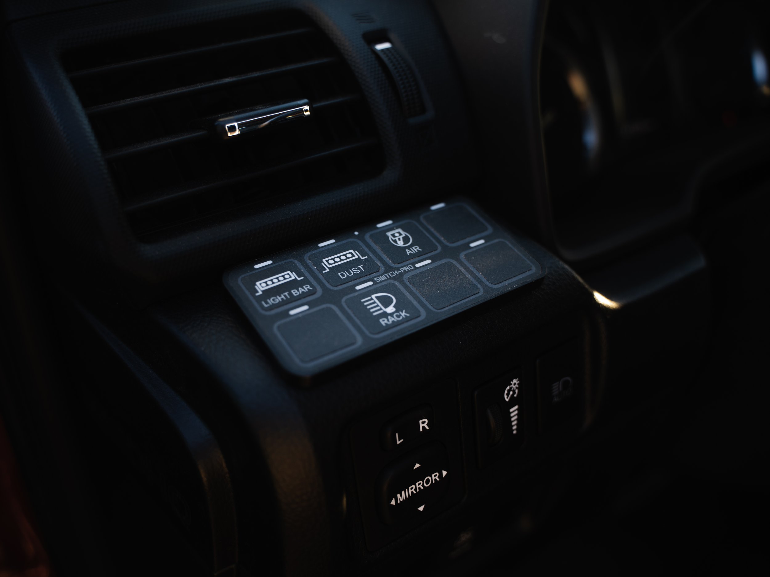 Close-up of a car interior dashboard with a control panel featuring buttons labeled 'Light Bar,' 'Dust,' 'Switch-Pro,' 'Air,' and a headlight icon, along with an air vent above.