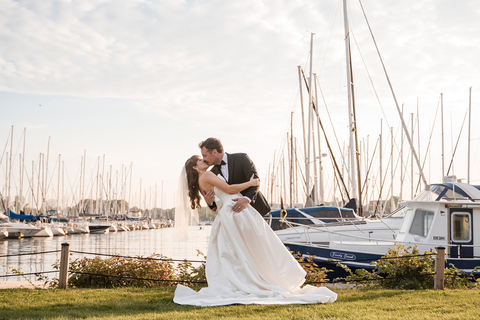 A bride and groom in wedding attire sharing a kiss and embracing in front of a marina with sailboats and a cloudy sky.