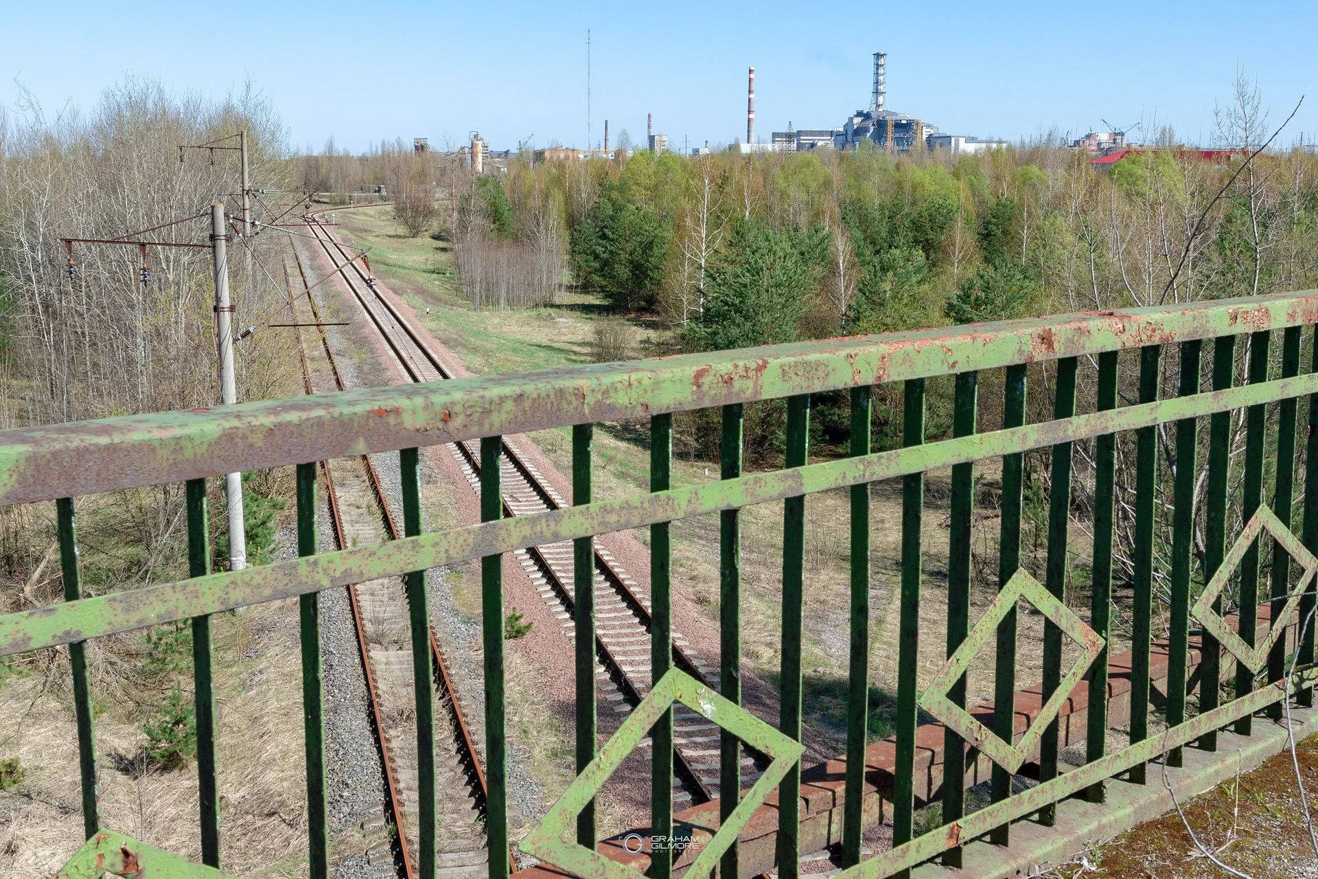 View from a bridge overlooking railroad tracks and a forest with industrial buildings in the background.
