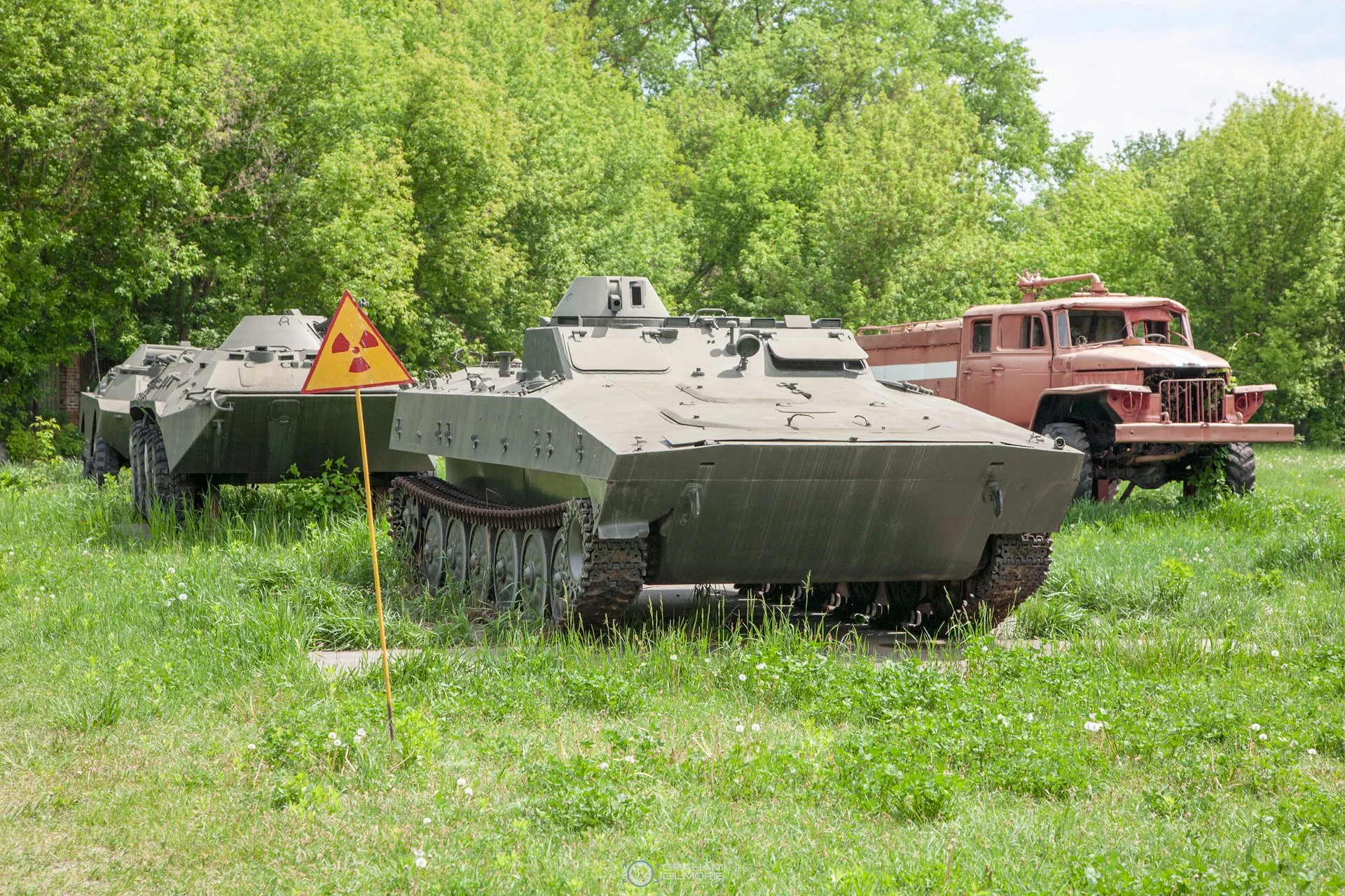 An outdoor display of military vehicles including an armored tank and an old truck, with a radioactive hazard sign in the grass.