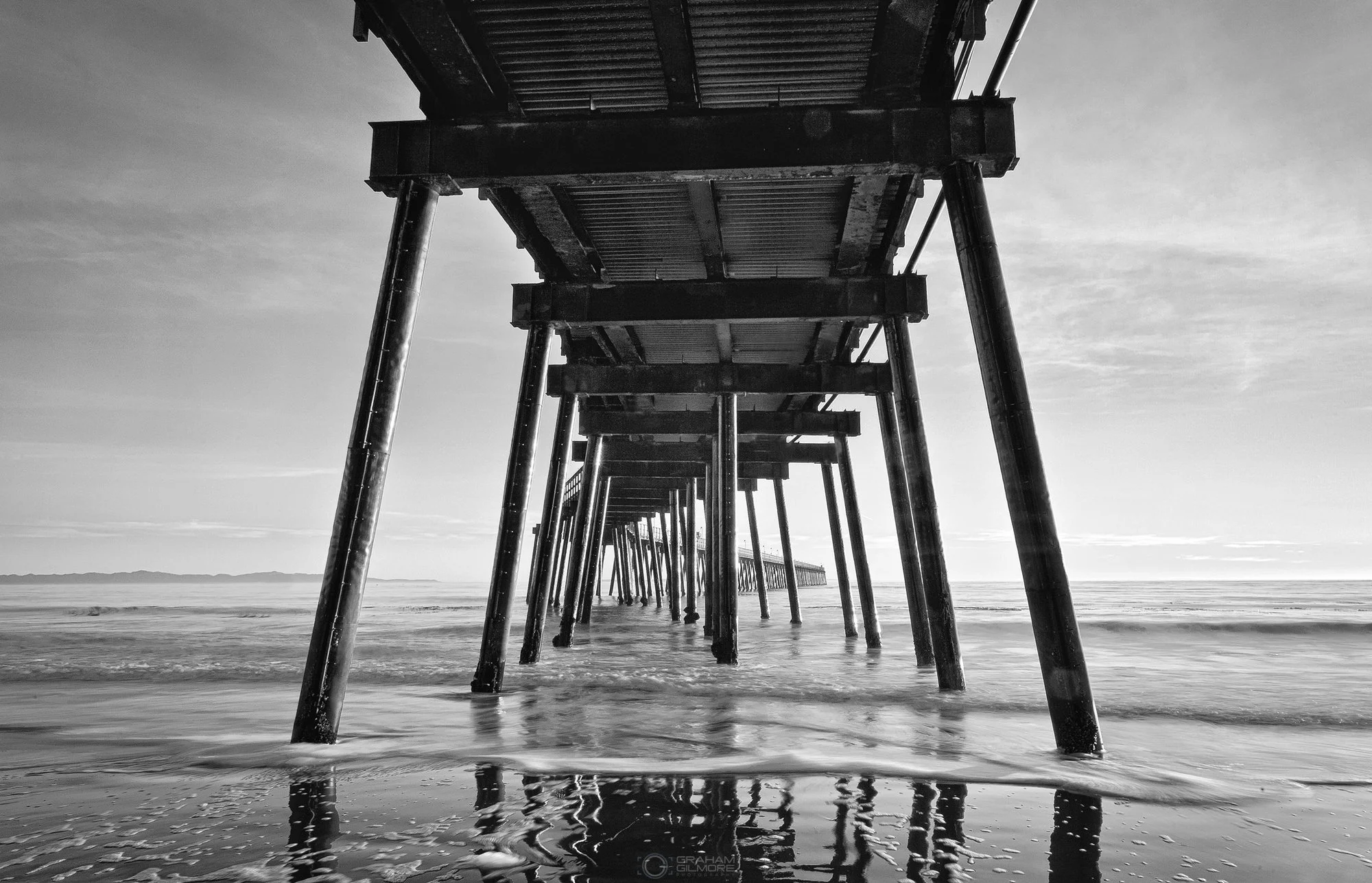 Black and white long exposure photograph of Ellwood Pier in Goleta, California, viewed from beneath with ocean waves moving around the columns and reflections on wet sand