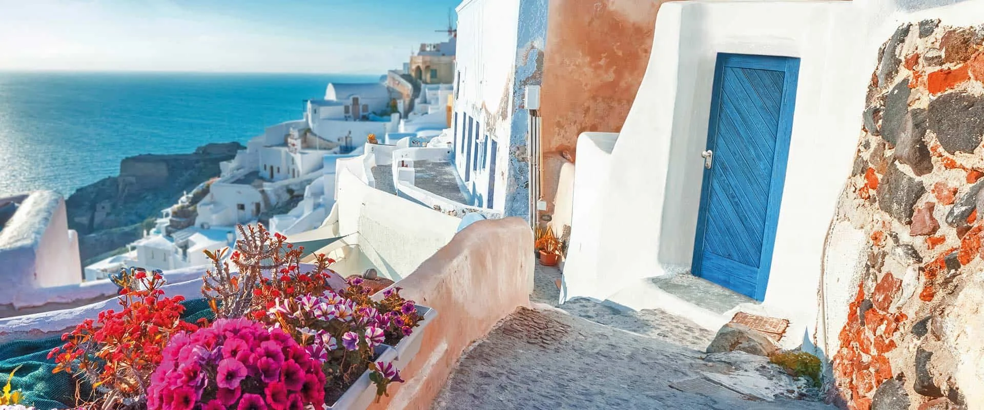 Veduta di case bianche a Santorini con il mare sullo sfondo, un cancelletto blu e piante e fiori colorati in primo piano