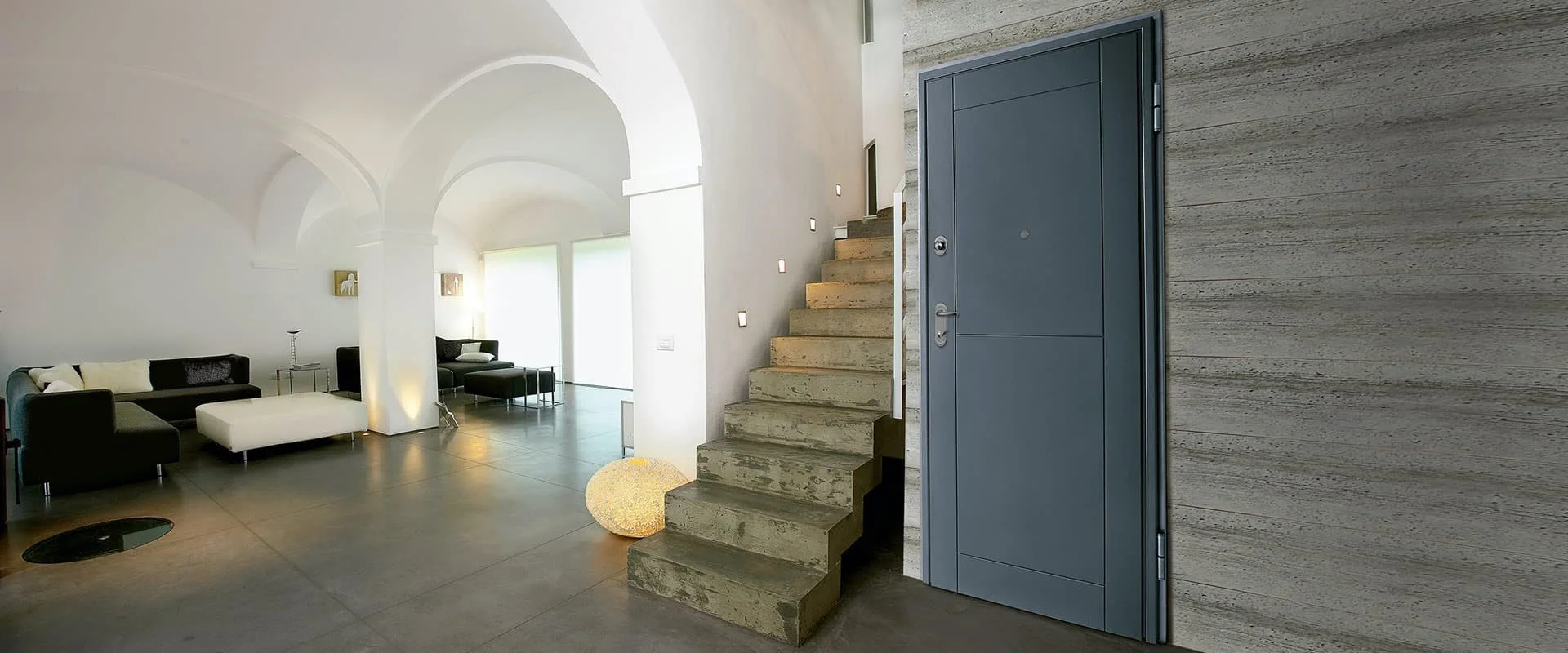 Interno di un modernissimo salotto con divani neri, una sedia e un tavolino, pareti bianche e grandi finestre, un'entrata con porta blu, un'elegante scala di cemento e pareti in soffice rivestimento.