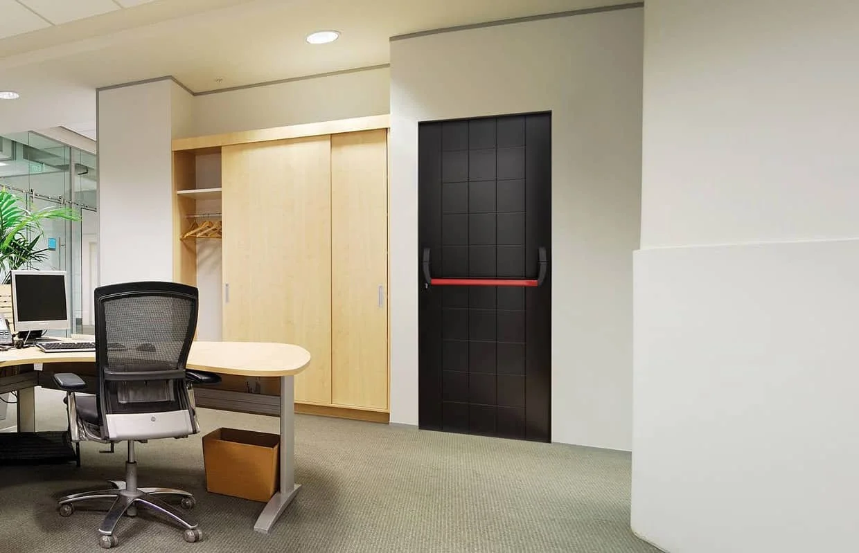 Ufficio con scrivania, sedia girevole e computer, armadio in legno chiaro, porta nera con maniglie, pareti bianche e pavimento in moquette. Obiettivo sul legno, porta e parte delle pareti dell'ufficio.