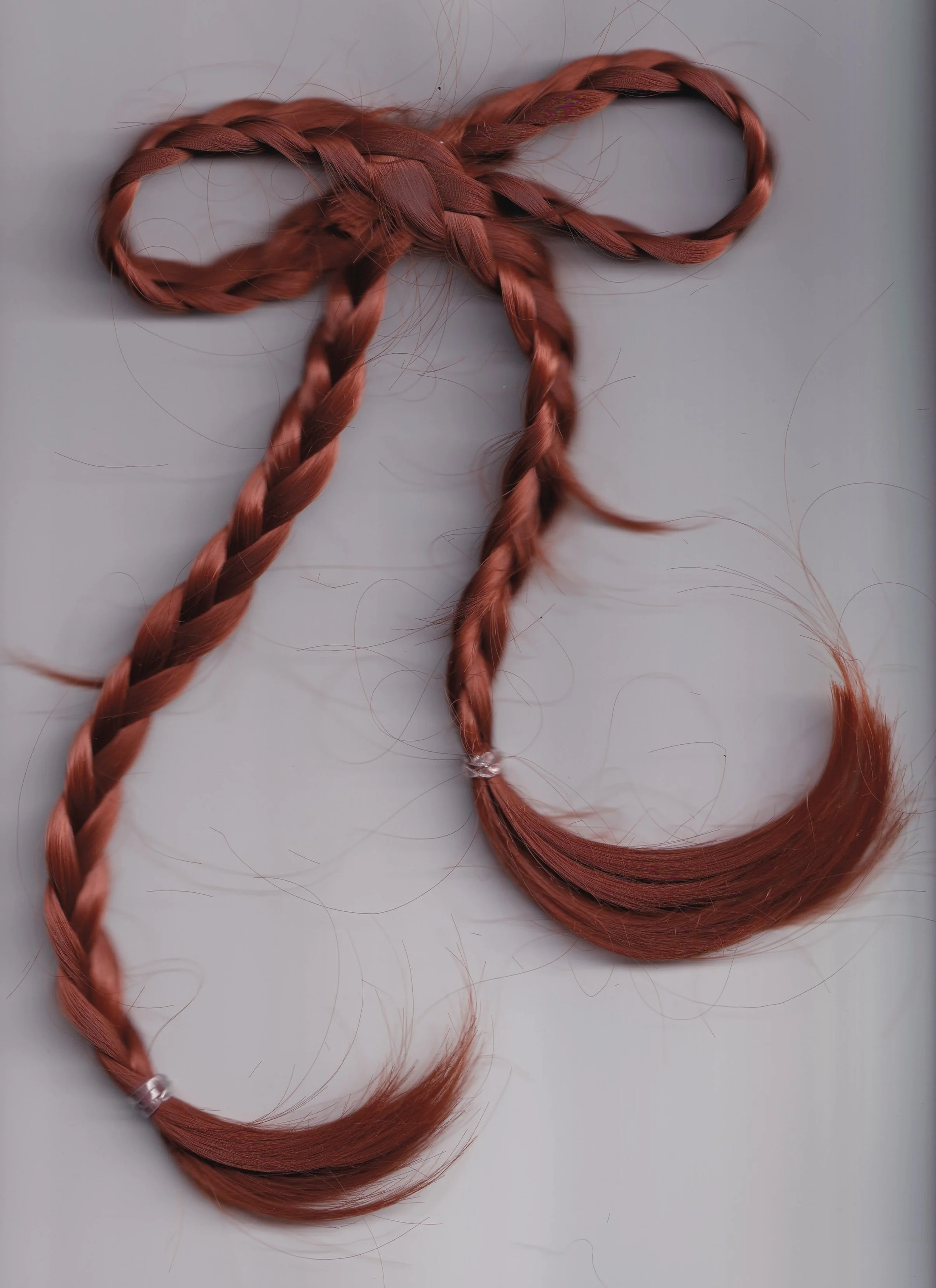 Two red hair braids, one with a loose end and the other tied with a clear hair tie, on a white surface.