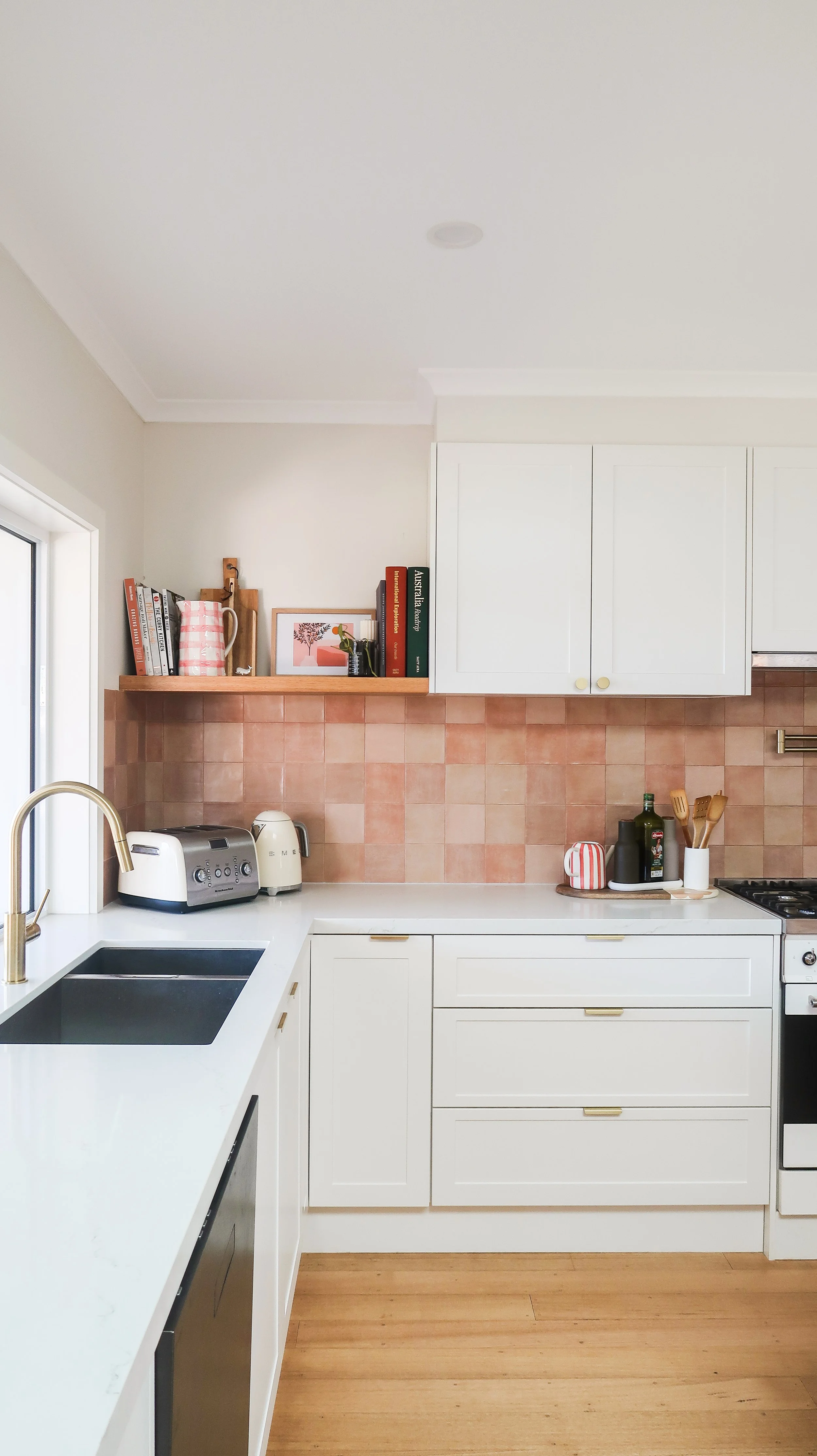 Kitchen with white cabinets, pink tile backsplash, wooden shelf with books and decor, black sink, toaster, kettle, oil bottles, and utensils.