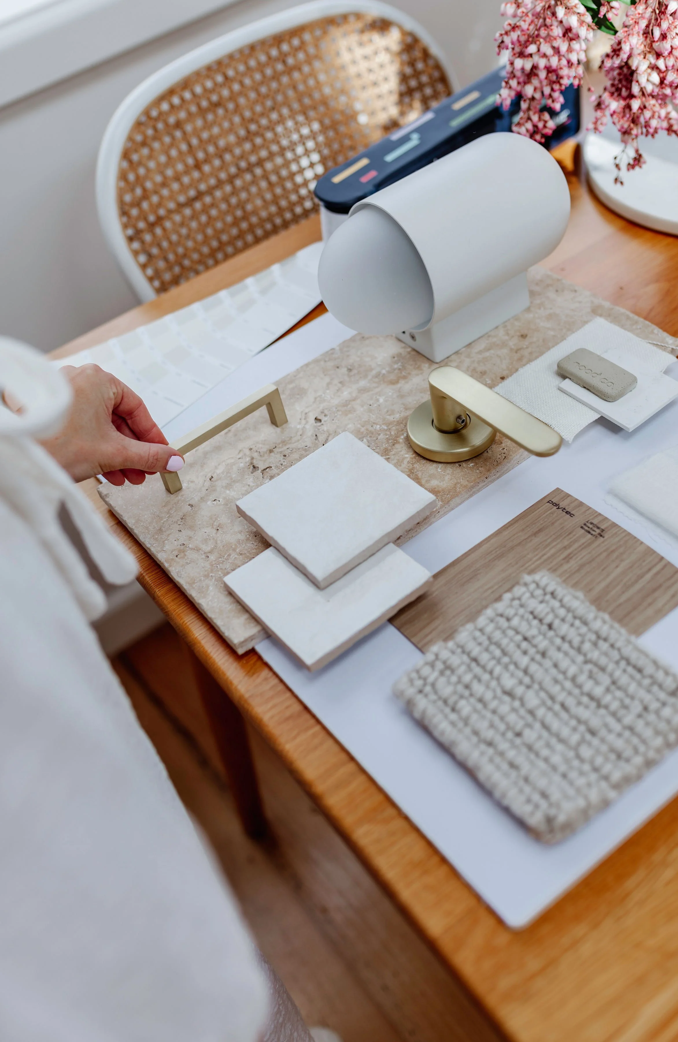 A table with various home decor materials, including tiles, fabric swatches, a paint color card, a small sample of a woven textile, a color palette, and a lamp, with a chair in the background.