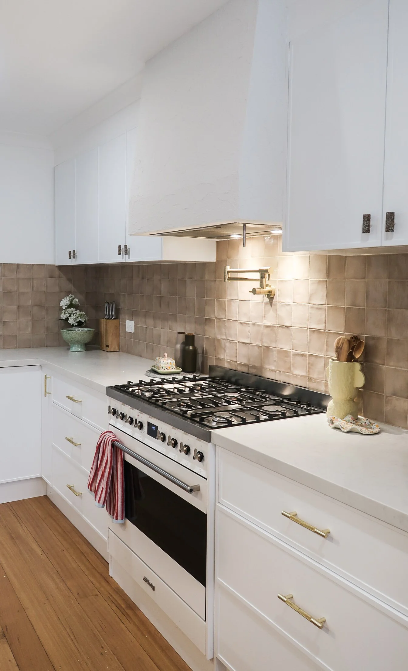 A modern kitchen with white cabinets, a beige tiled backsplash, and a white oven with a black stovetop. Decorative items include a vase with flowers, a wooden knife block, and a yellow pottery container with wooden spoons. The kitchen floor is wood.