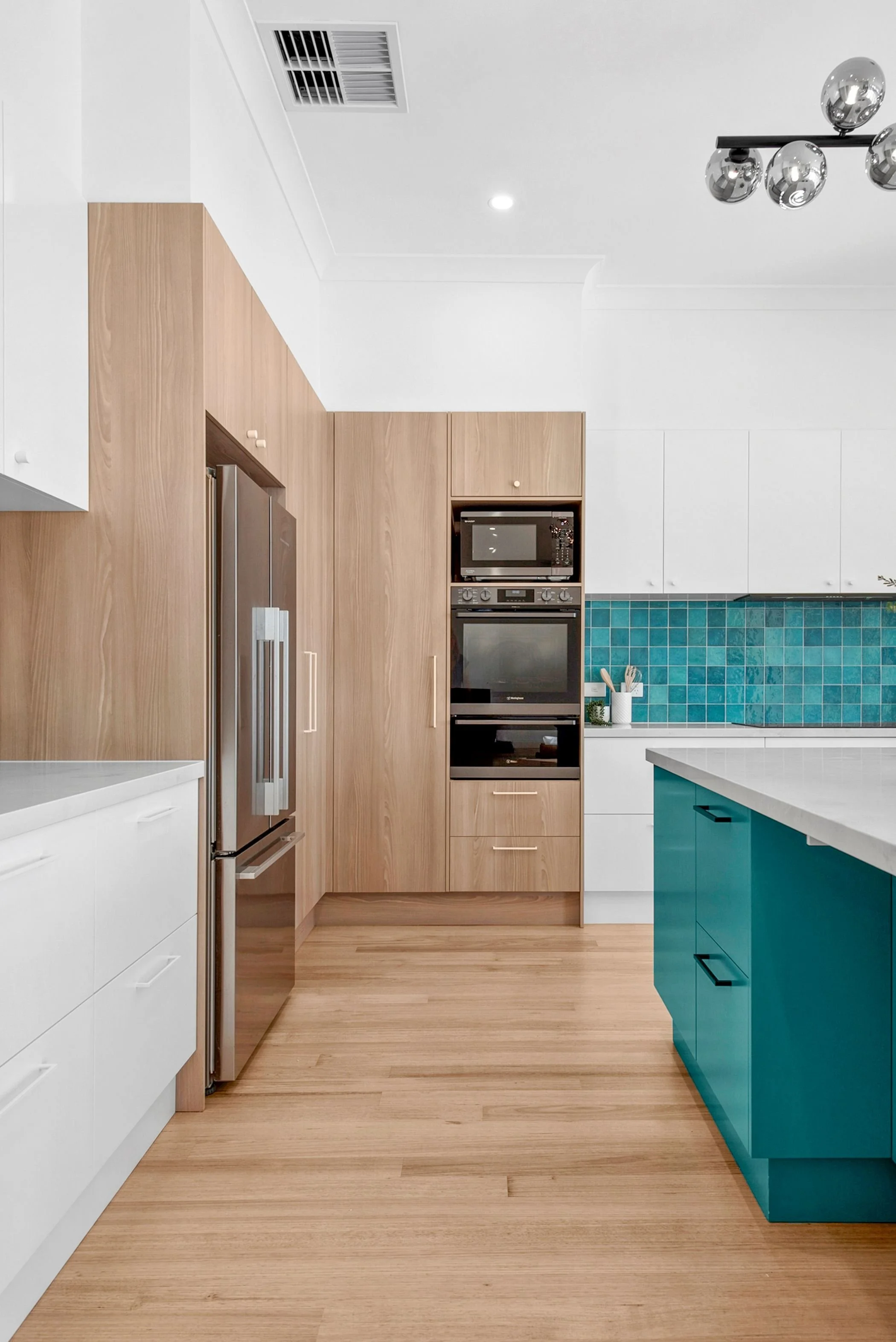 Modern kitchen with white and wood cabinetry, a green-tiled backsplash, a kitchen island with a marble countertop and three wooden barstools, stainless steel refrigerator, oven, and gas stove, wooden flooring, and contemporary lighting fixture.