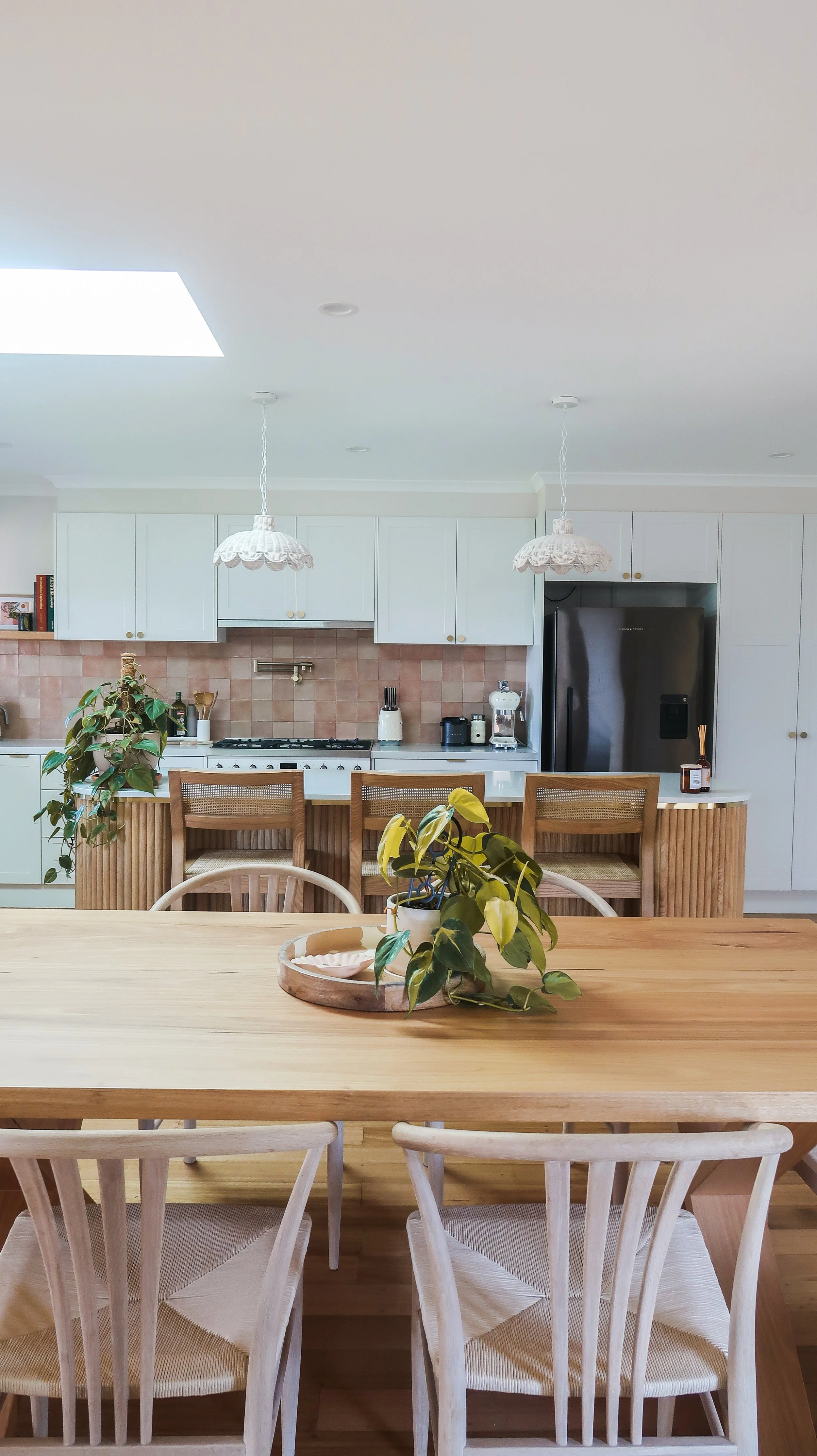 A modern kitchen with white cabinets, pink tile backsplash, black appliances, and a wooden dining table with chairs. Potted plants are on the table and near the island, with pendant lights hanging from the ceiling.