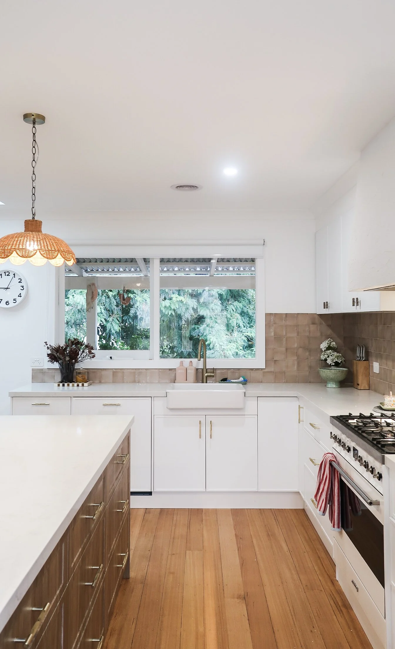 Modern kitchen with white cabinets, wooden flooring, and a large window overlooking greenery. Contains a white farmhouse sink, gold faucet, gas stove, and decorative plants.