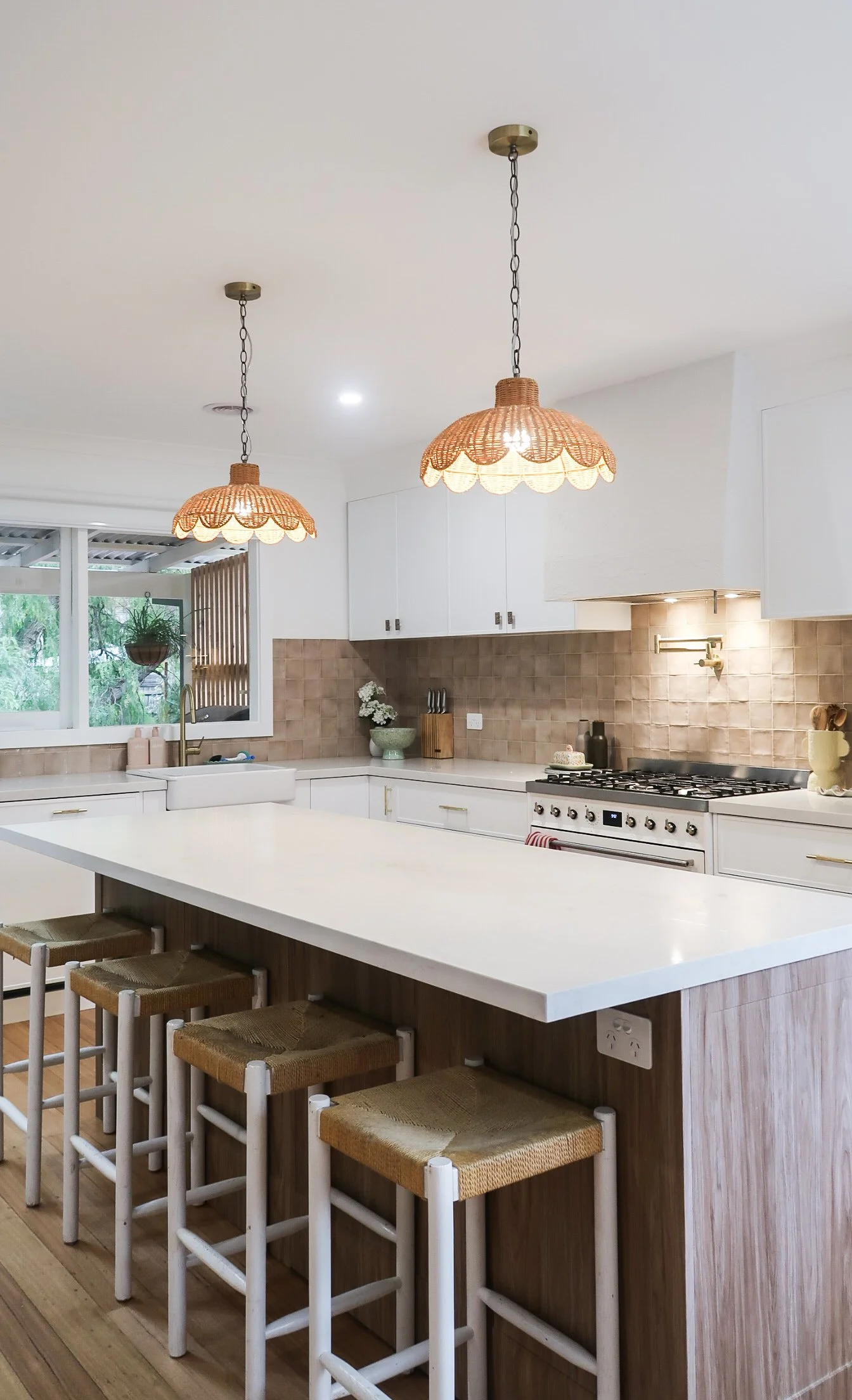 A modern kitchen with white cabinets, a beige tile backsplash, a white farmhouse sink, a white island with four wooden stools, gold fixtures, pendant lights with woven shades, and a window overlooking greenery.