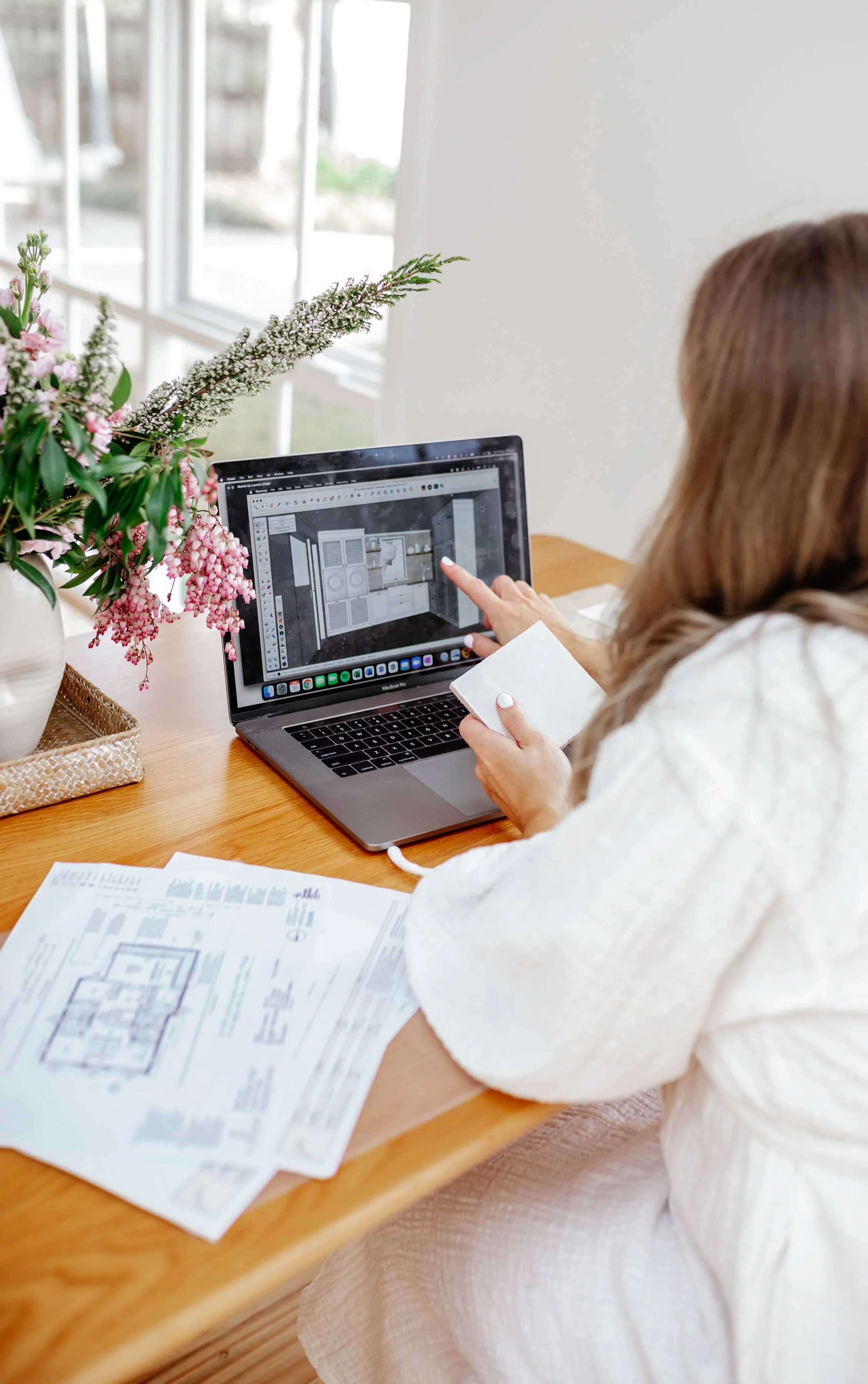 A woman working on a laptop at a wooden table, looking at a digital interior design of a room on the screen, with architectural plans spread on the table, next to a vase with pink and white flowers.