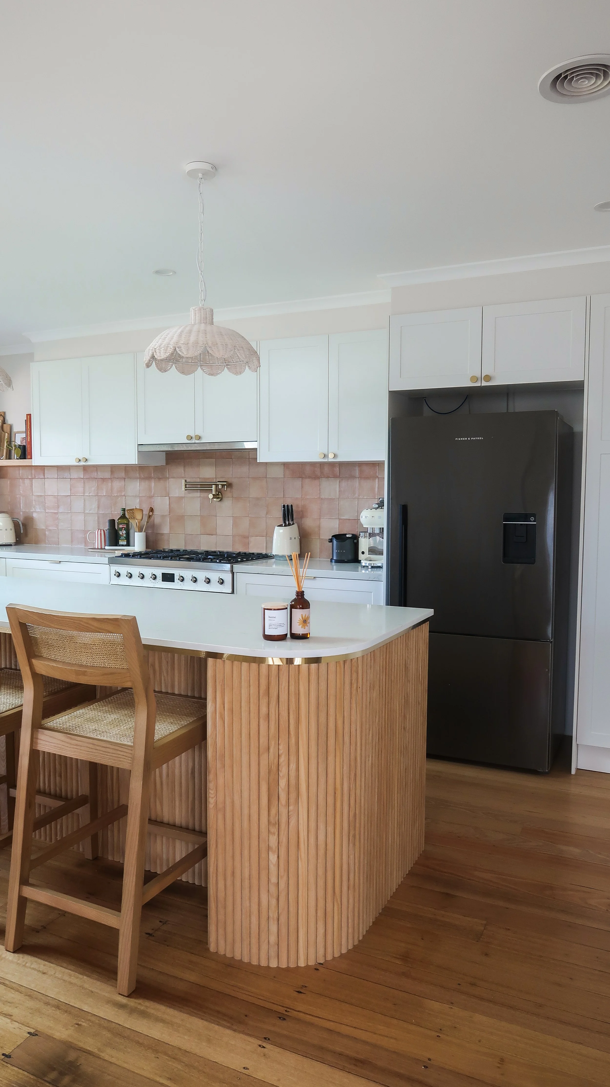 A kitchen with white cabinets, a black refrigerator, a white countertop, and a curved wooden kitchen island with two chairs. There is a pink backsplash, various small appliances, and a pendant light with a scalloped shade.
