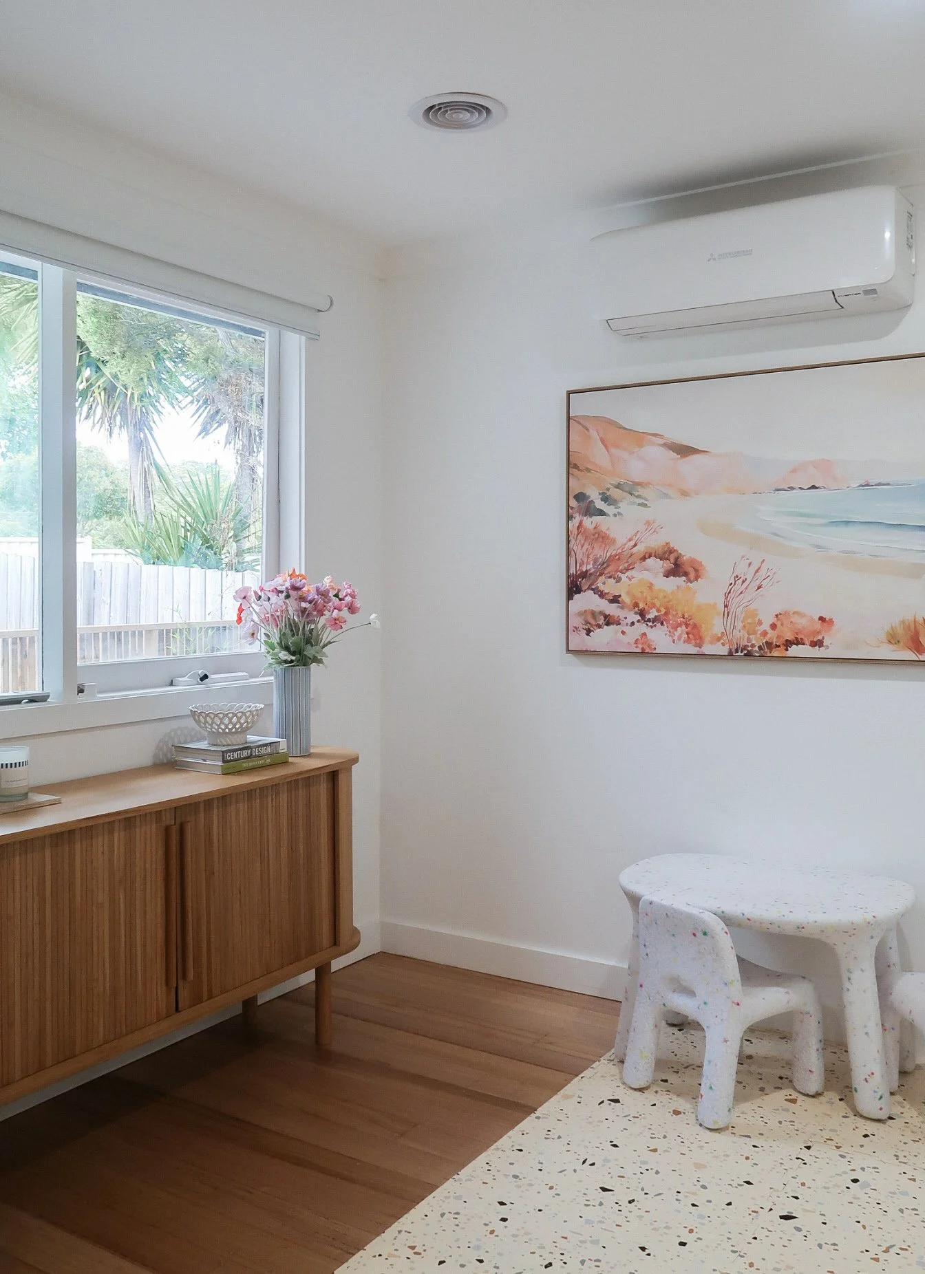 A cozy room with a wooden sideboard, a bouquet of pink and white flowers in a vase, a window with a view of trees and a fence, a colorful coastal landscape painting on the wall, and a speckled white children's table with chairs.