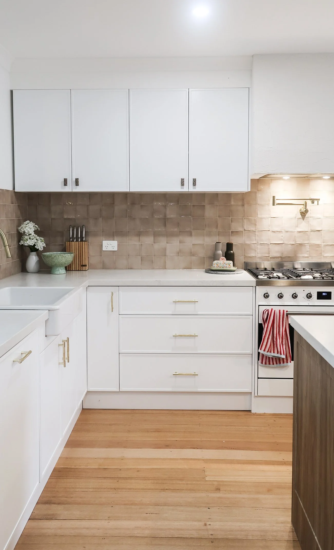 A modern kitchen with white cabinets, beige tiled backsplash, a stainless steel stove with an oven, and a wooden floor. Decor includes a vase with white flowers, a green ceramic bowl, a knife block, and vases on the countertop, as well as a red and w