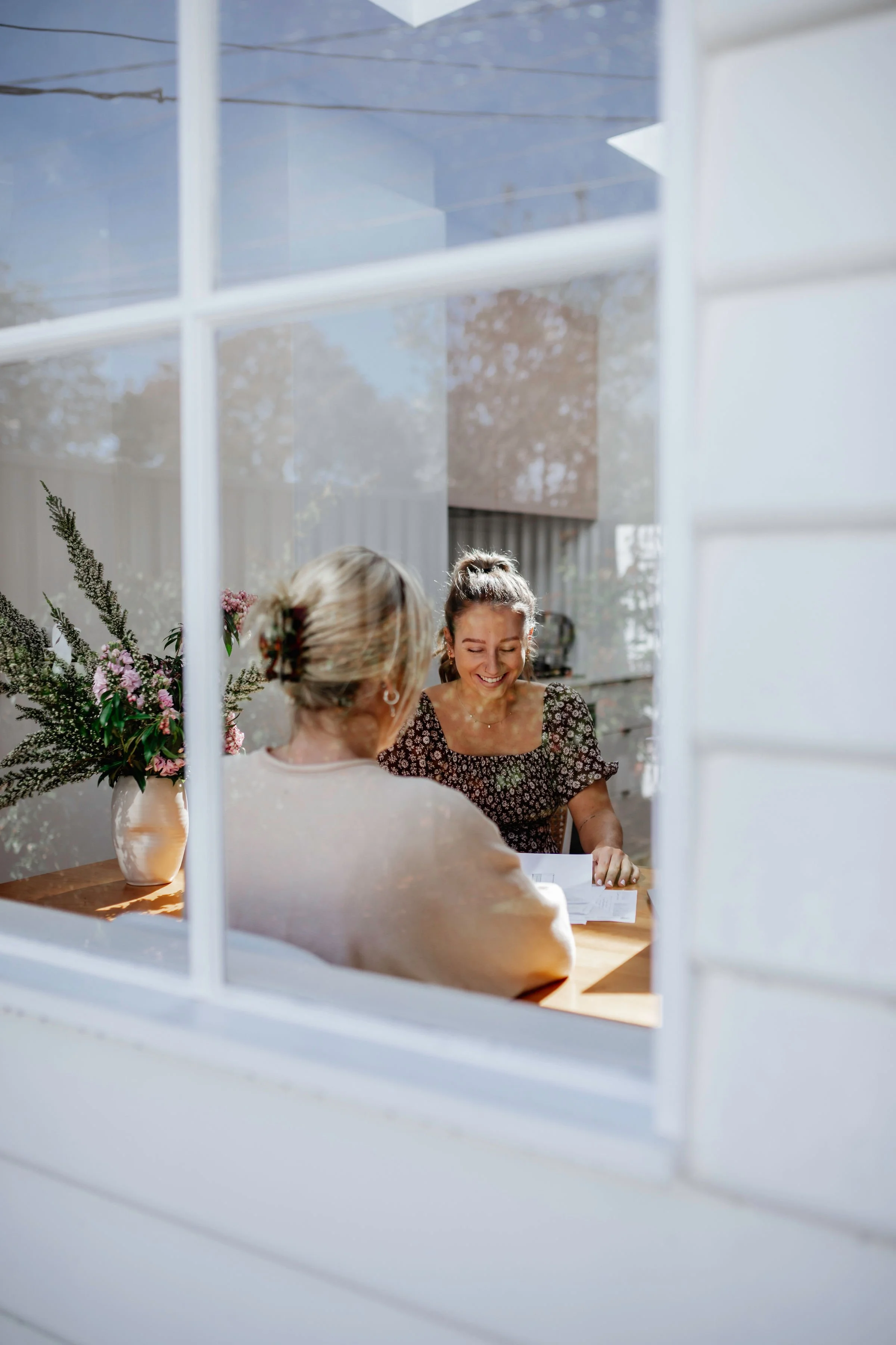 Two women are sitting at a table by a window, smiling and talking. The table has a vase with pink and purple flowers, and sunlight is streaming through the window.
