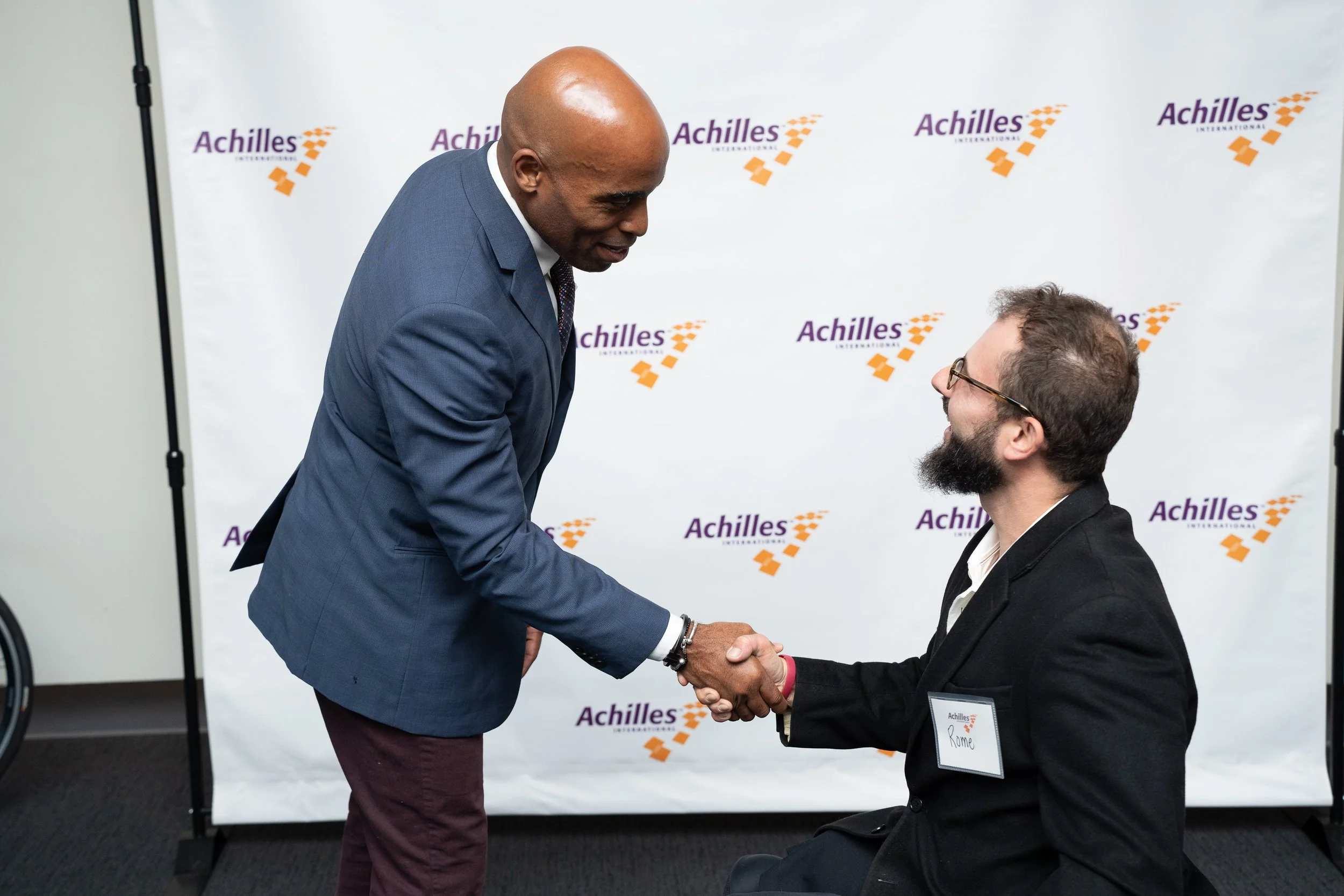  Tiki Barber shakes athlete’s hand in wheelchair in front of Achilles branded step and repeat 